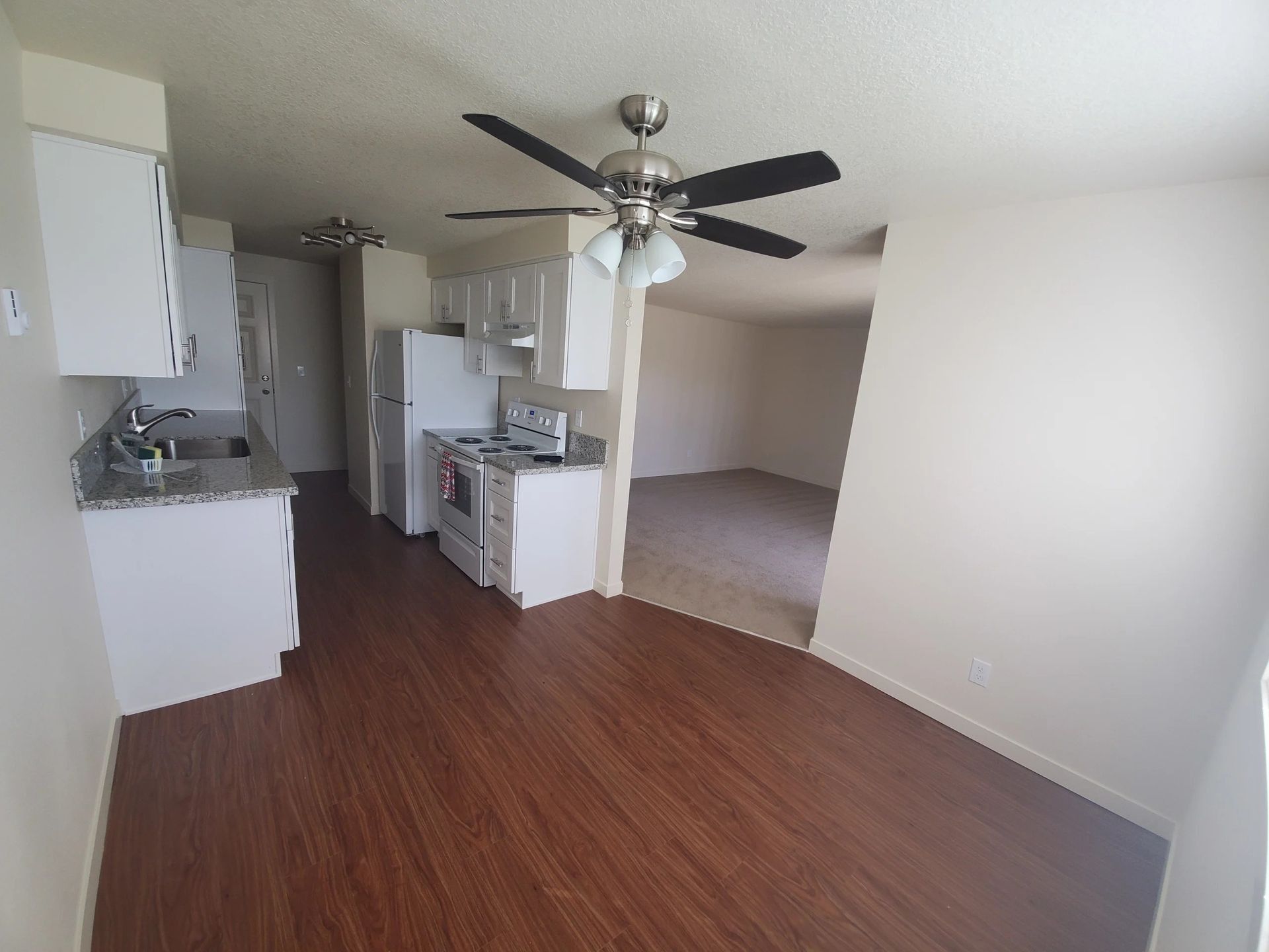 Empty apartment living area with wood floors, ceiling fan, kitchen, and bedroom alcove visible.