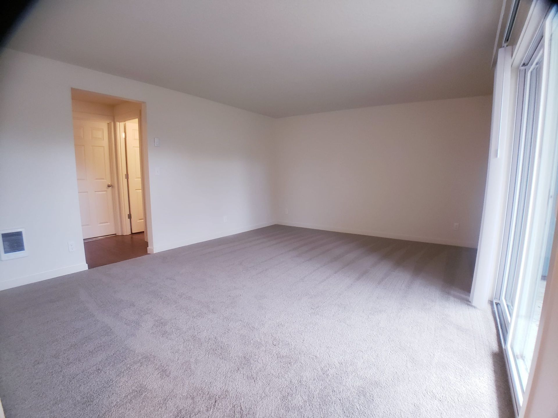Empty beige living room with carpet, open doorway, and sliding glass door to the right