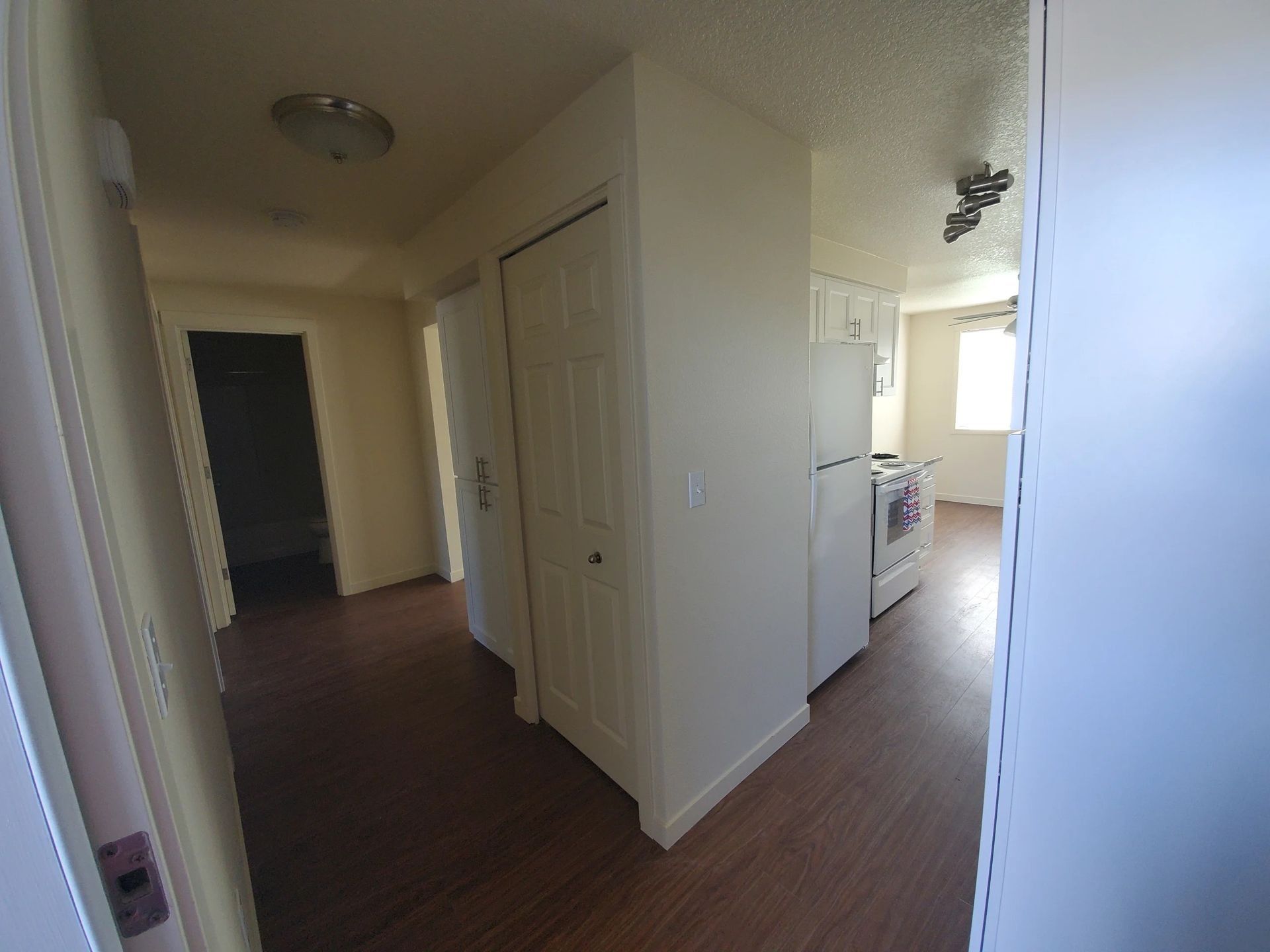 Hallway and kitchen area in a home with white walls, dark wood floors, and appliances visible.