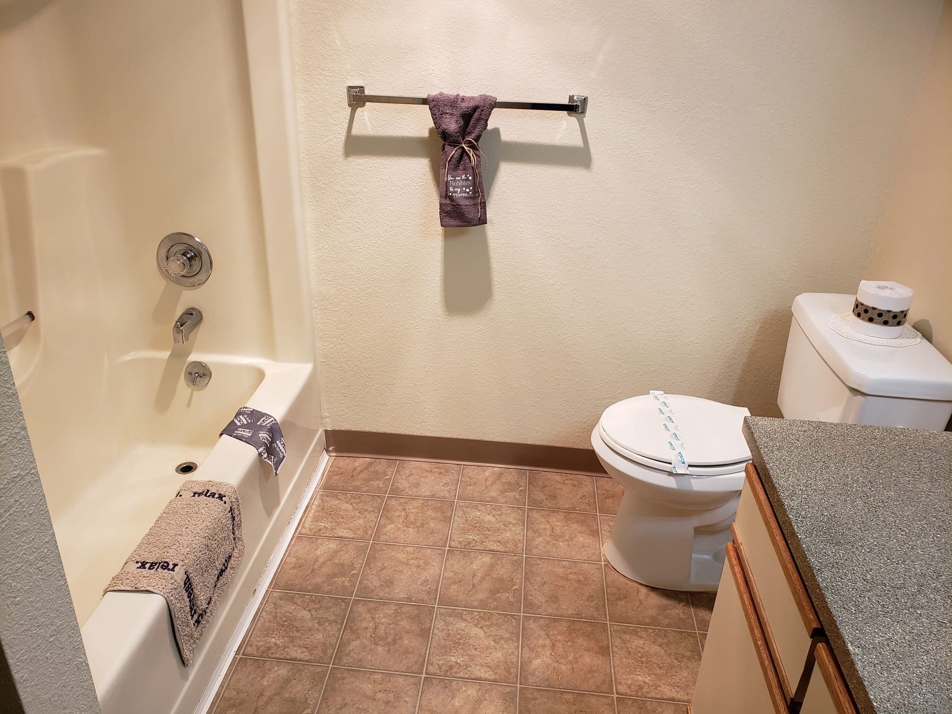 Small bathroom with bathtub, toilet, and granite counter, featuring towels on racks and tan tile floor.