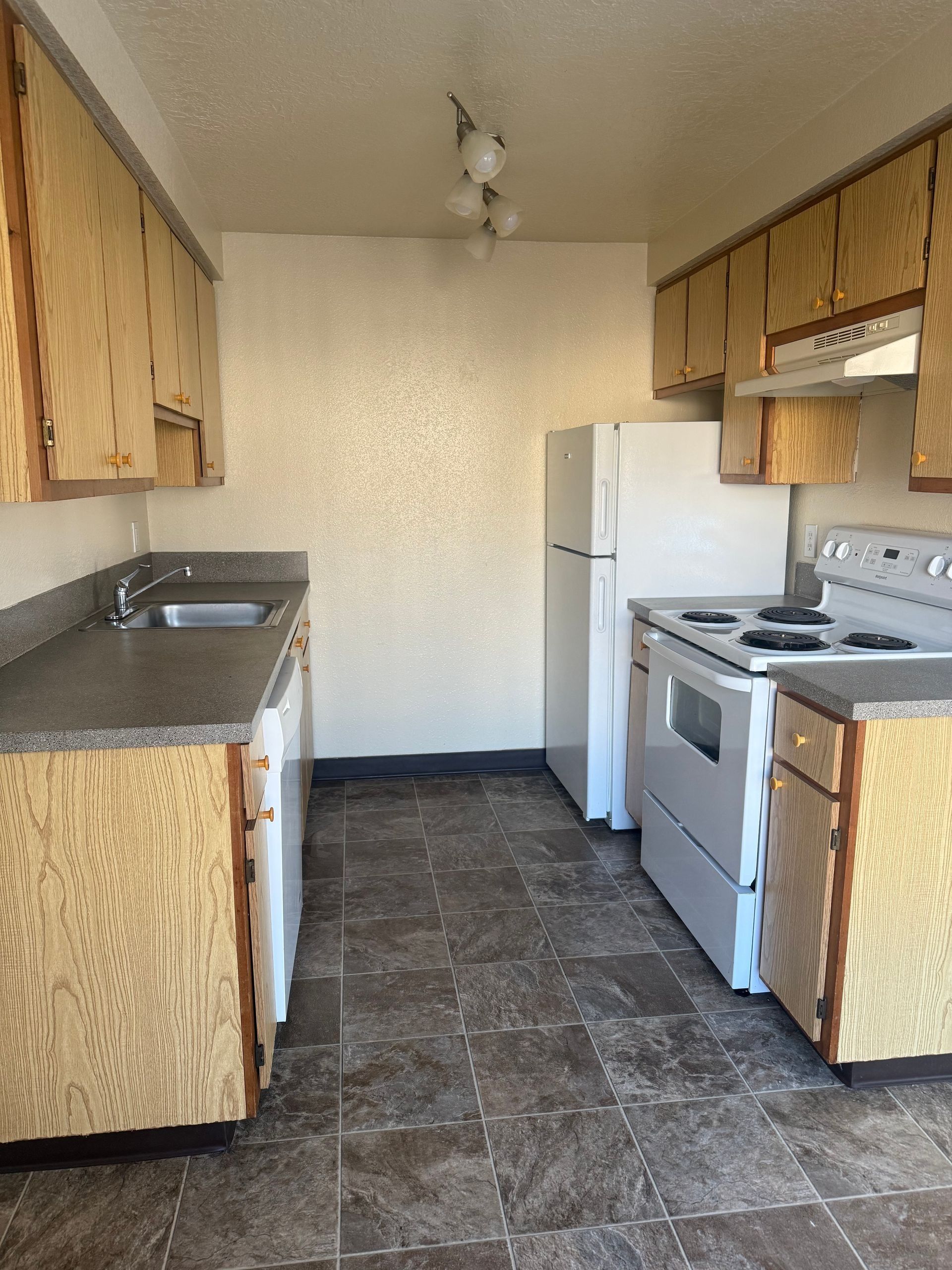 Narrow vintage kitchen with wood cabinets, white appliances, and tiled floor.