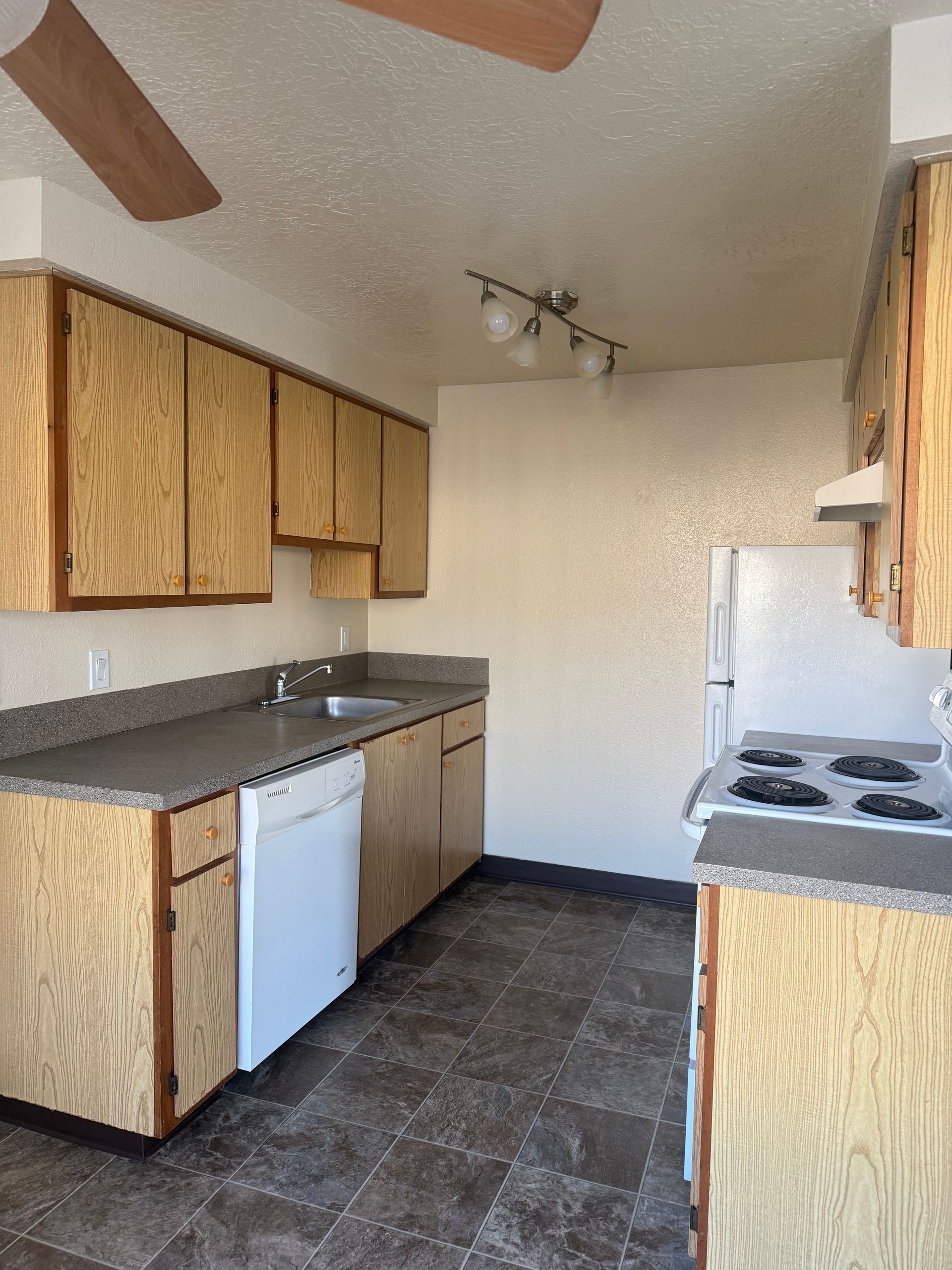 Small kitchen with wood cabinets, dark countertops, white appliances, and a ceiling fan