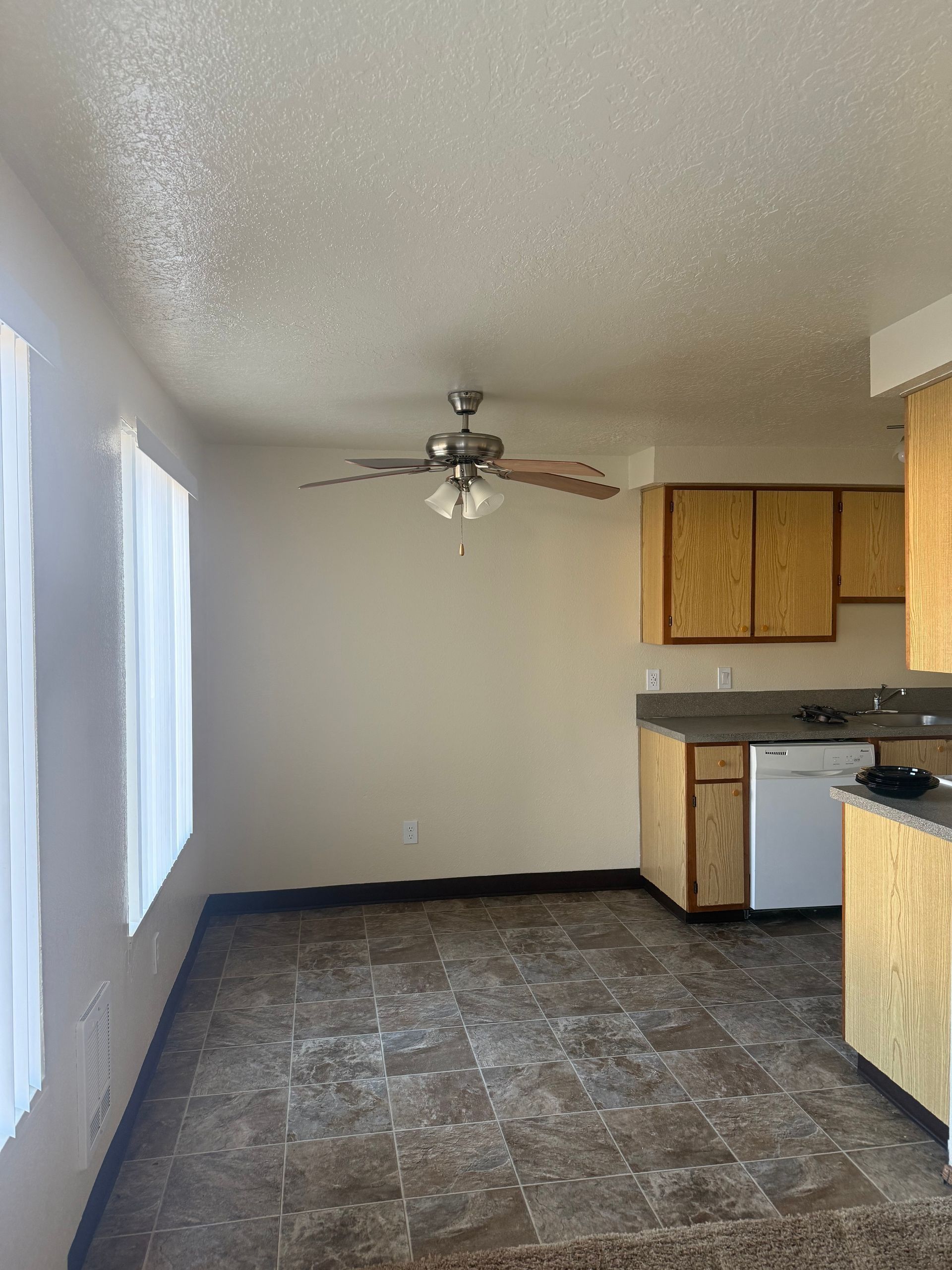 Empty kitchen with gray tile floor, white walls, ceiling fan, and wood cabinets.