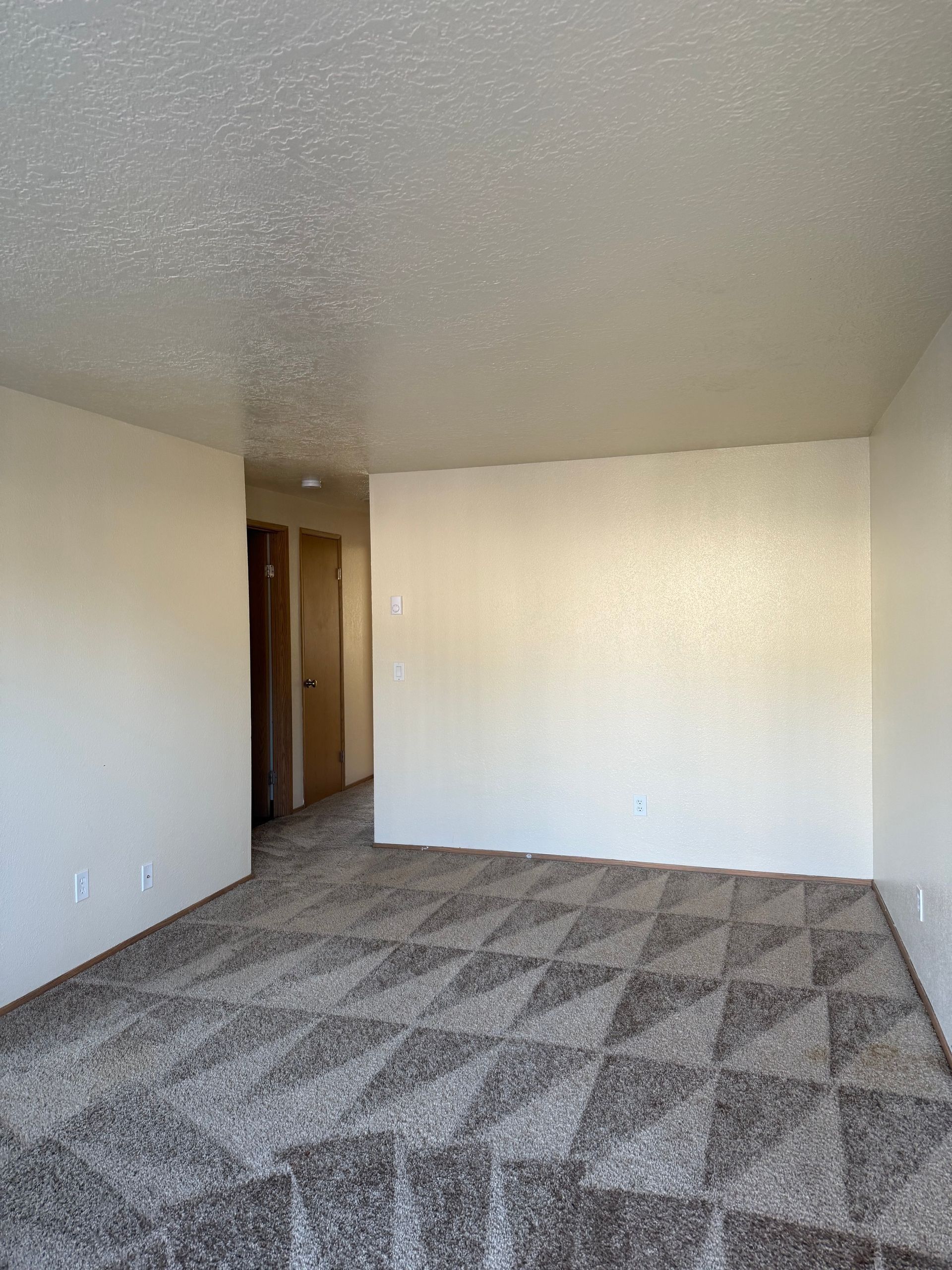 Empty beige carpeted room with textured ceiling and an open doorway to a hallway.