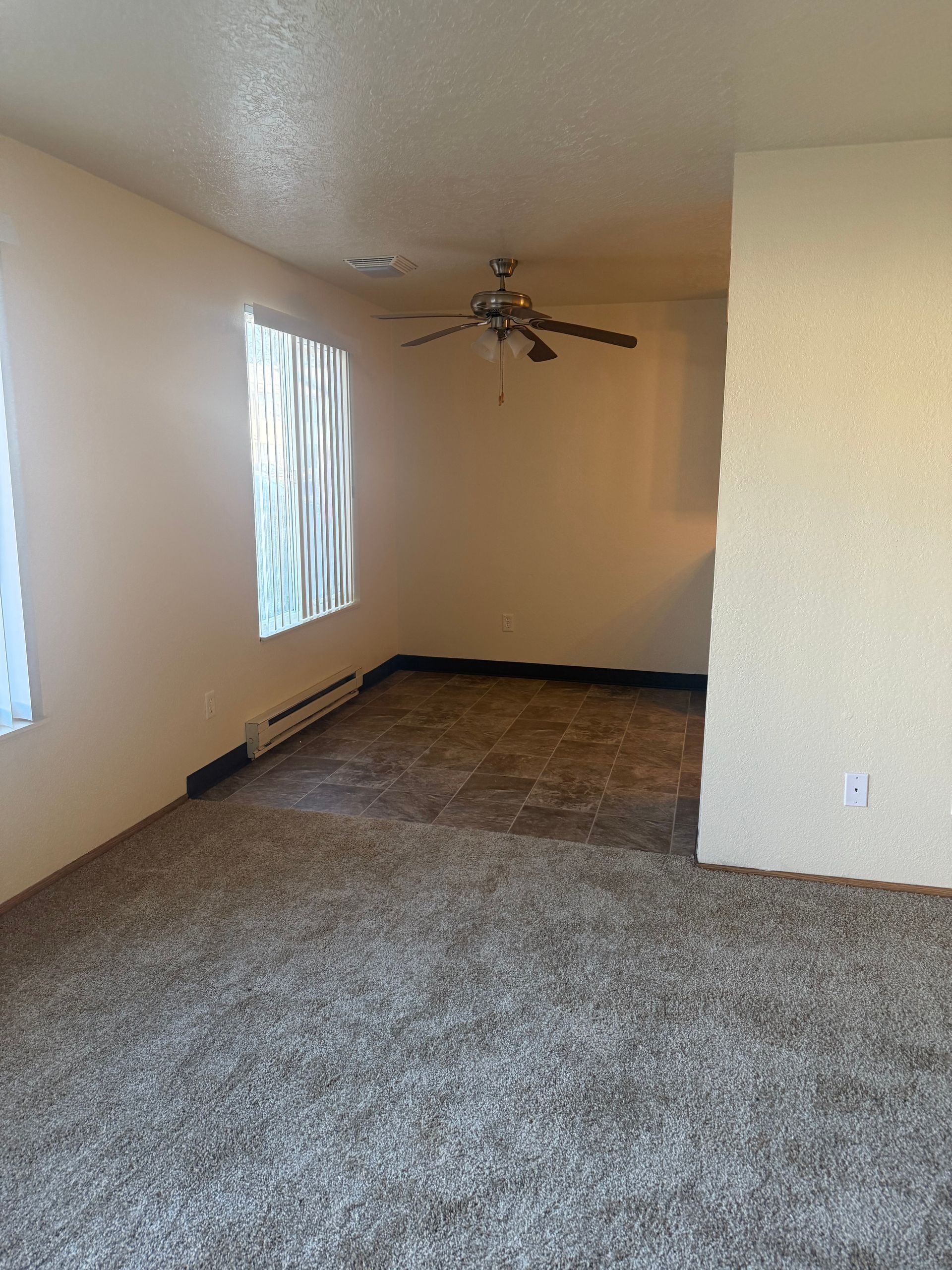 Empty carpeted living room with beige walls, two windows, and a ceiling fan