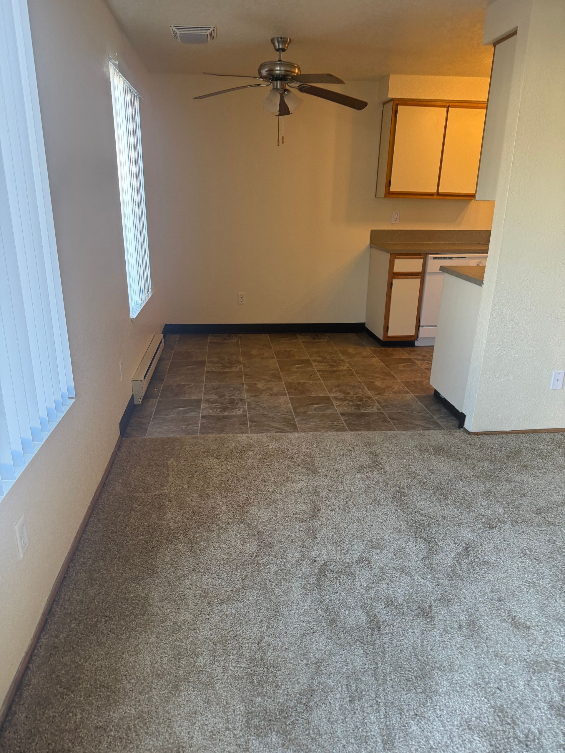 Empty apartment living area with carpet, ceiling fan, and small kitchen at the back