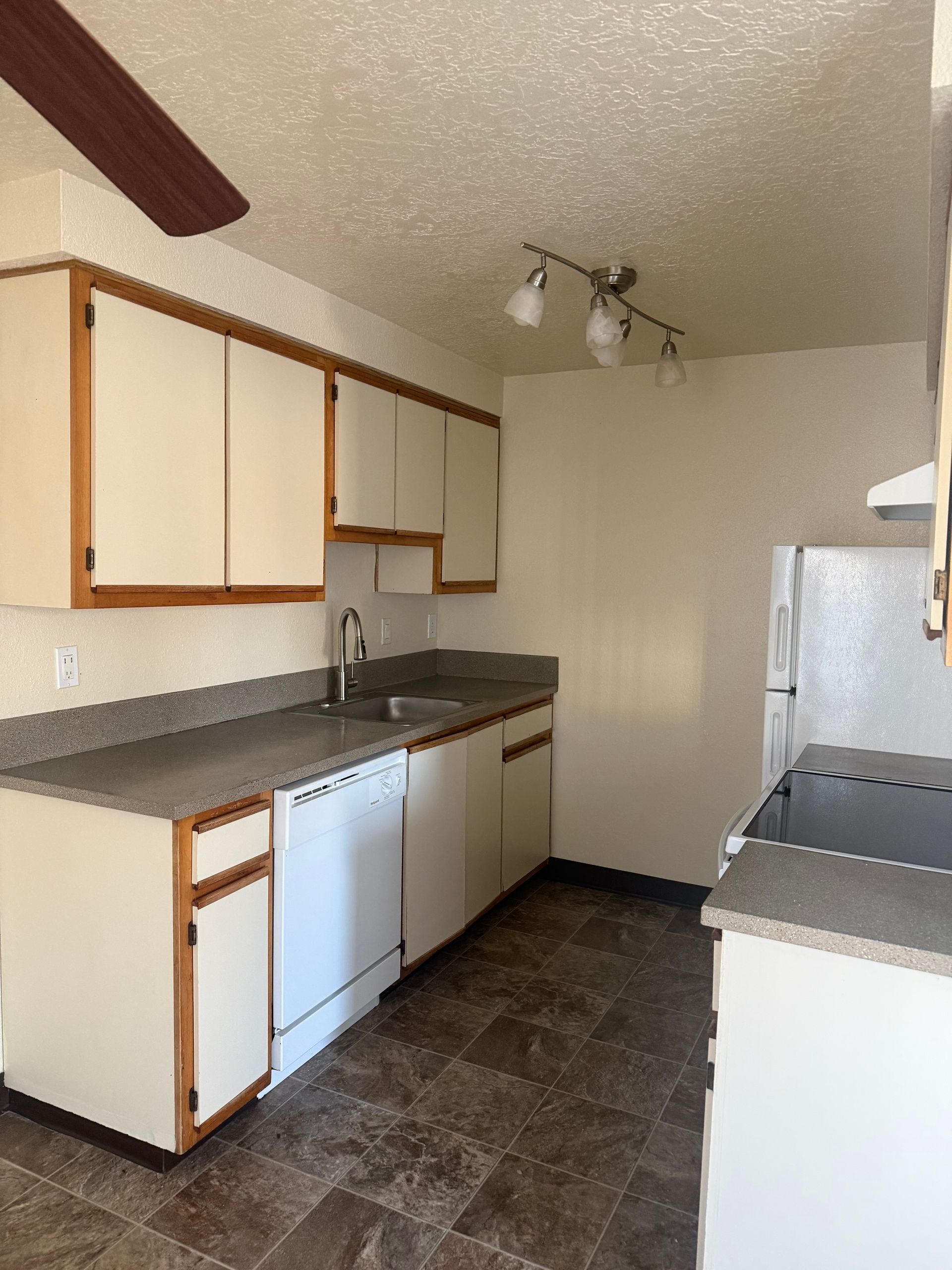 Empty kitchen with white cabinets, dark countertops, and tiled floor under track lighting.