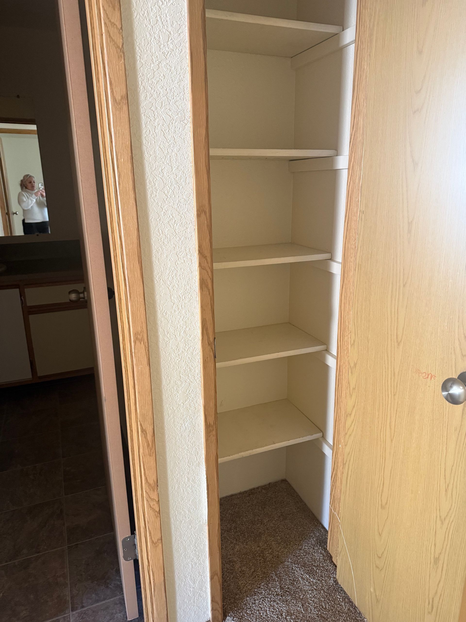 Narrow white closet with empty shelves and carpeted floor, seen through an open wooden door.