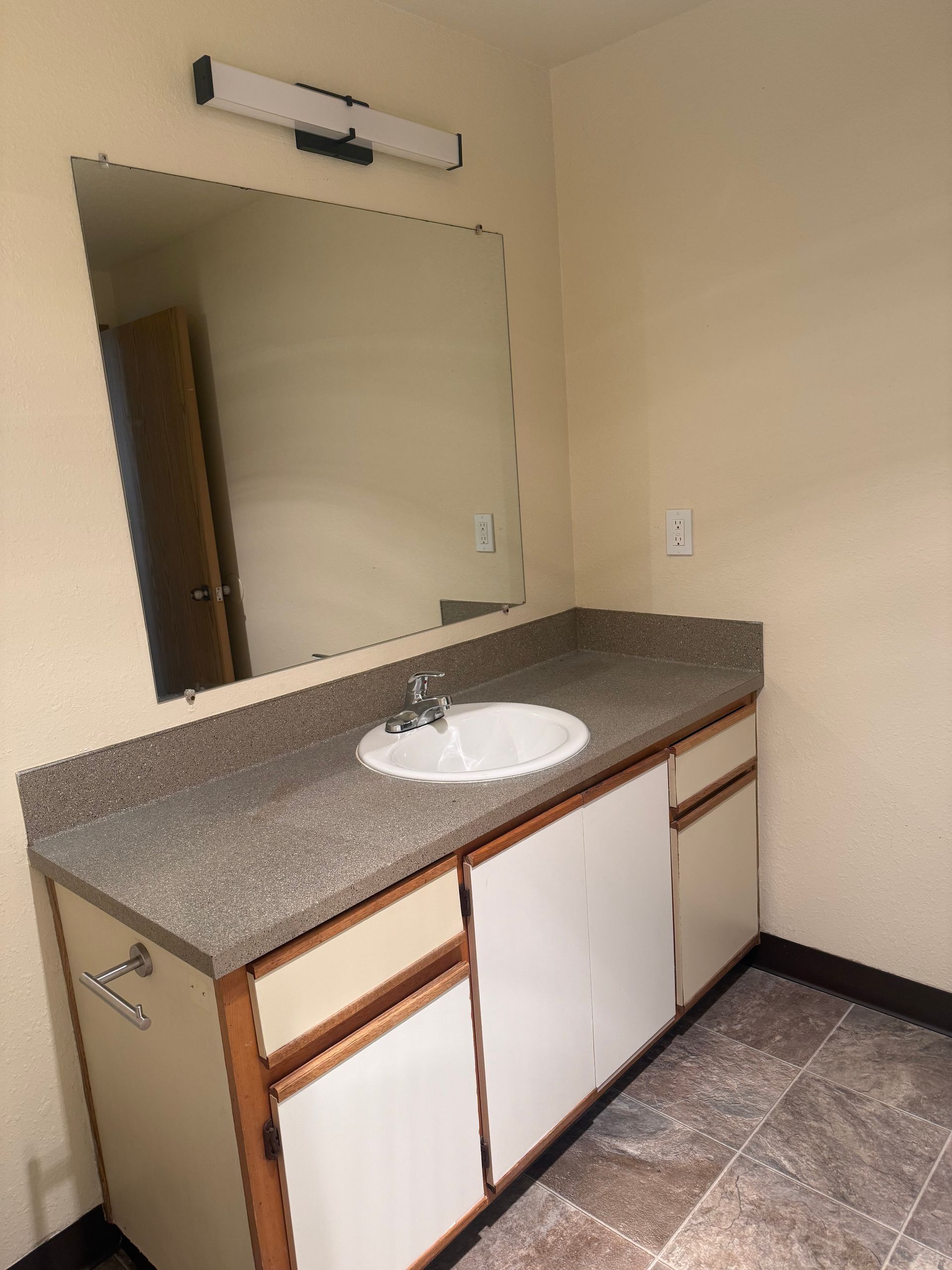Bathroom vanity with sink, speckled countertop, large mirror, and wall light in a beige room