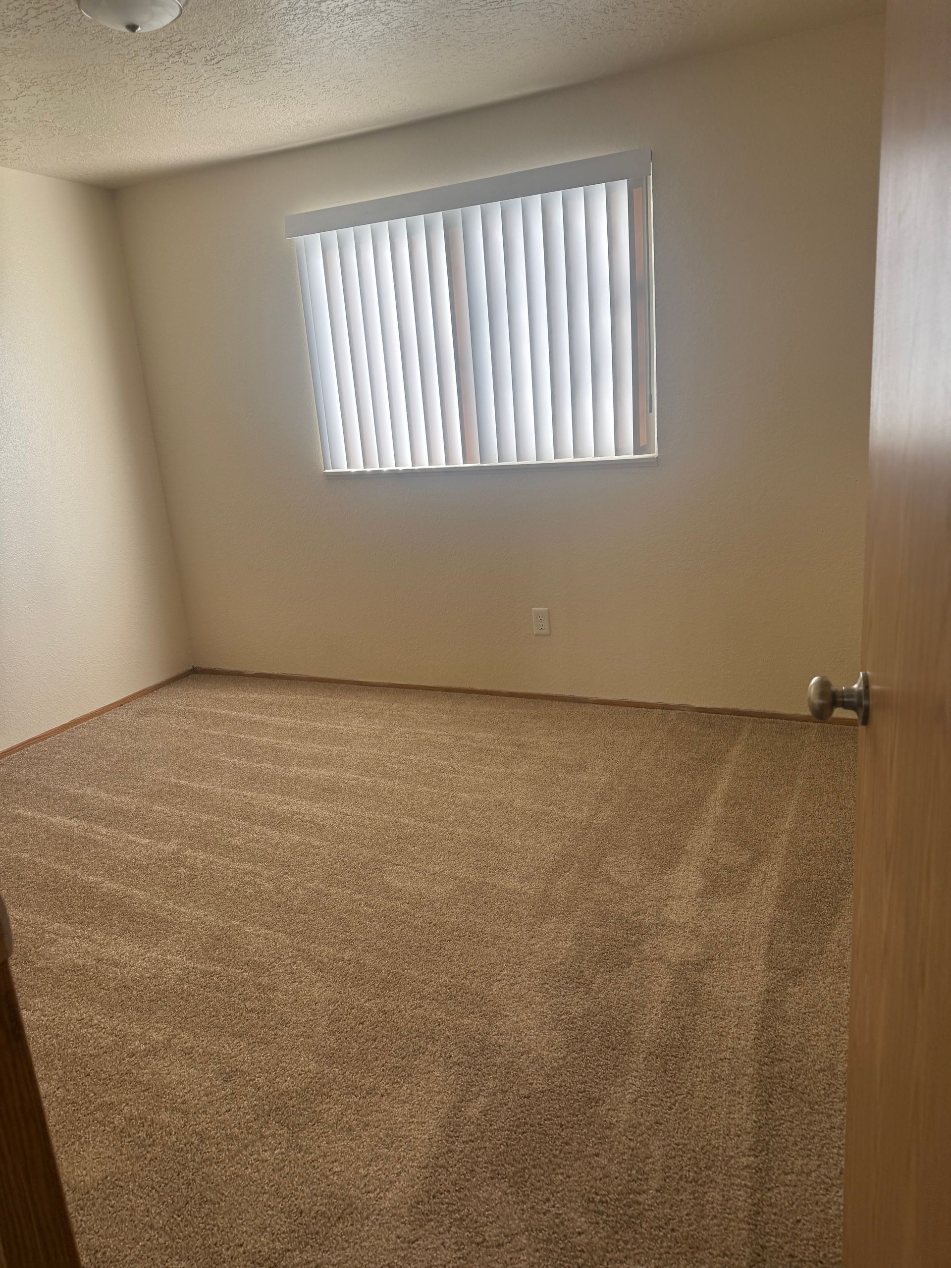Empty carpeted room with beige walls and a small window with vertical blinds