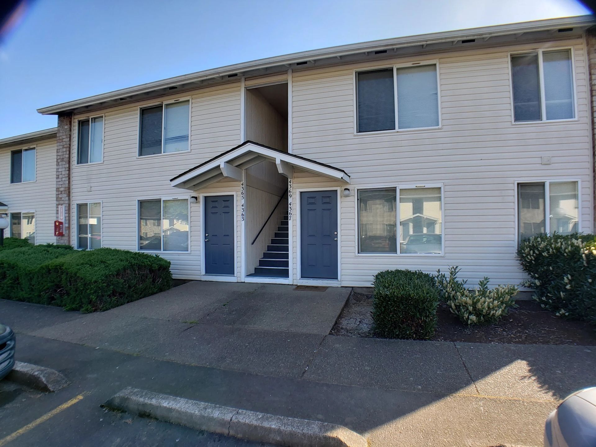 Two-story beige apartment building with blue doors, white trim, and bushes in front