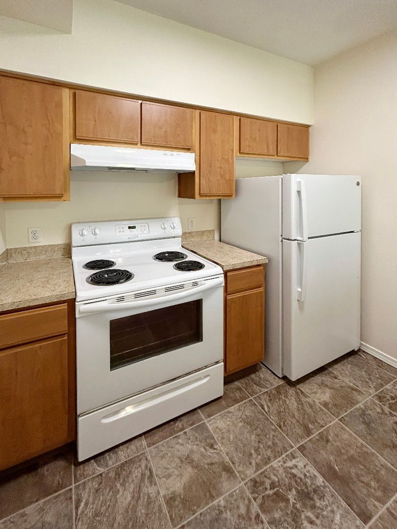 Small kitchen with wood cabinets, white stove, countertop, and white refrigerator on tile floor.