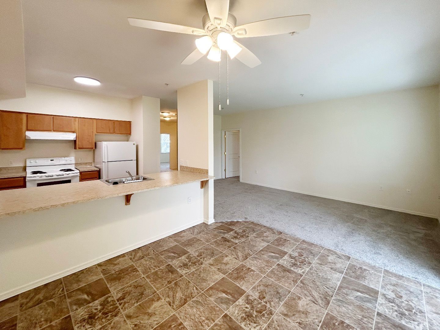 Empty open-plan apartment with kitchen pass-through, white ceiling fan, and carpeted living area