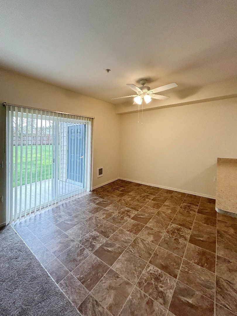 Empty tiled living room with sliding glass door, ceiling fan, and beige walls.