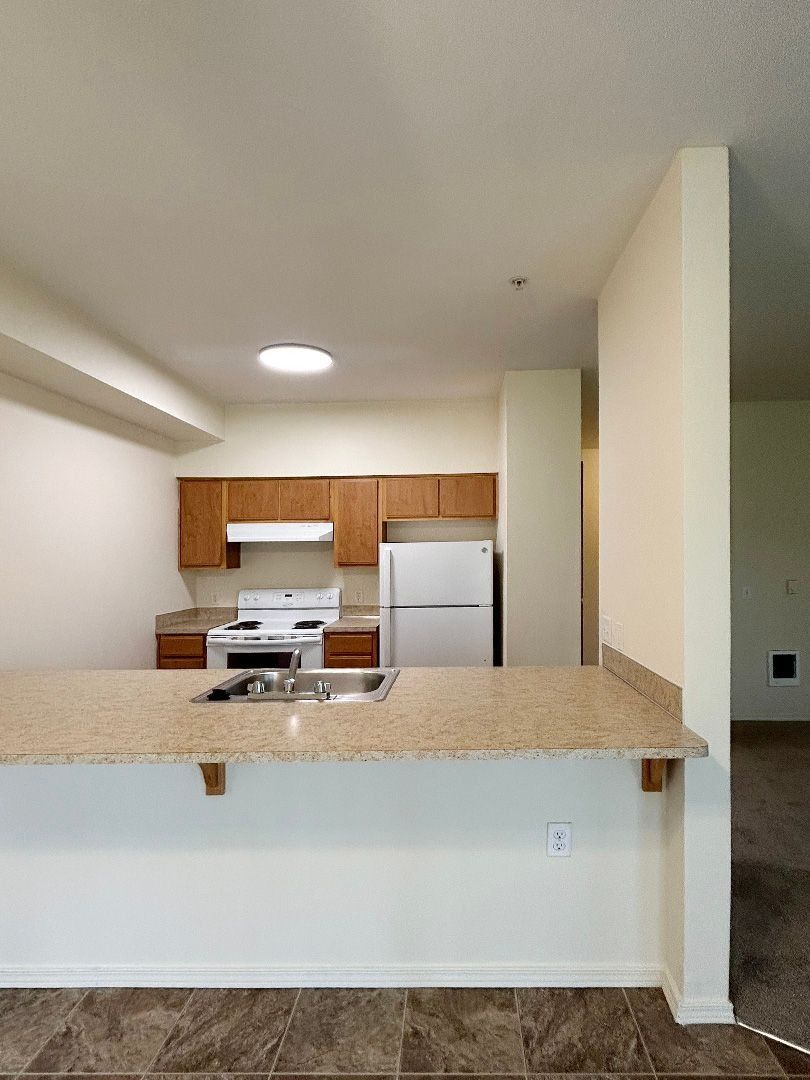 Empty kitchen with white appliances, wood cabinets, and a granite breakfast bar in an open apartment.