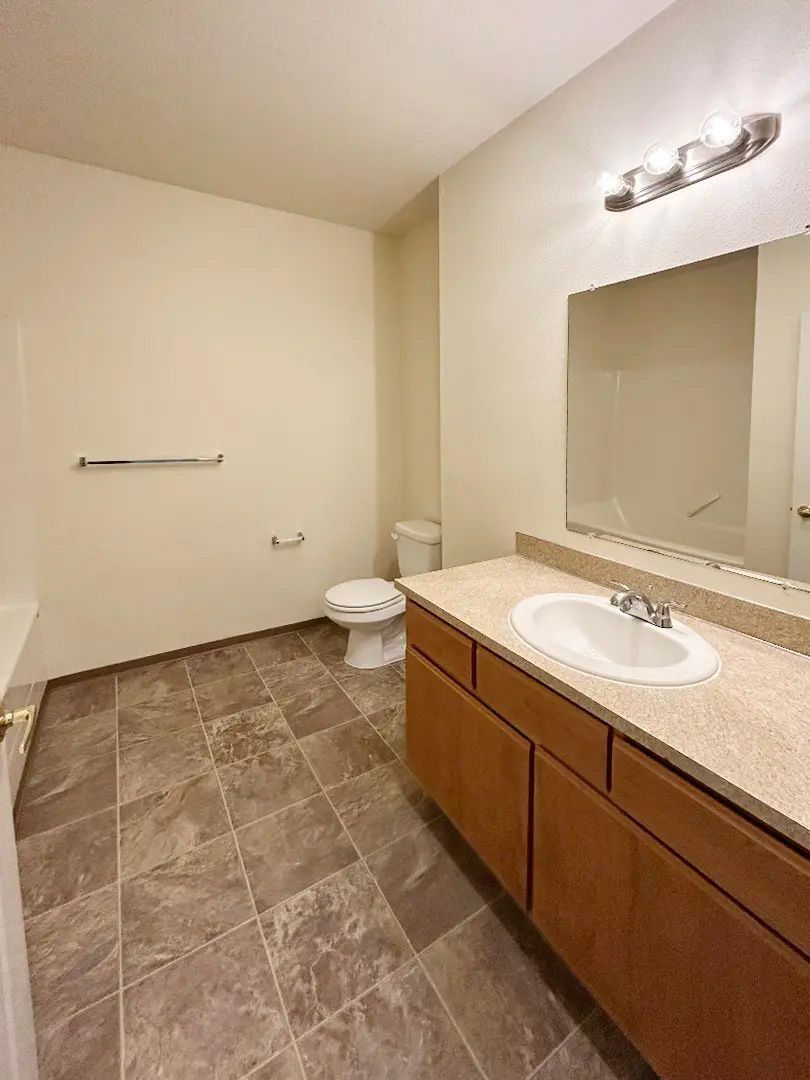Empty bathroom with beige tile floors, wooden vanity, large mirror, toilet, and wall-mounted towel rack.