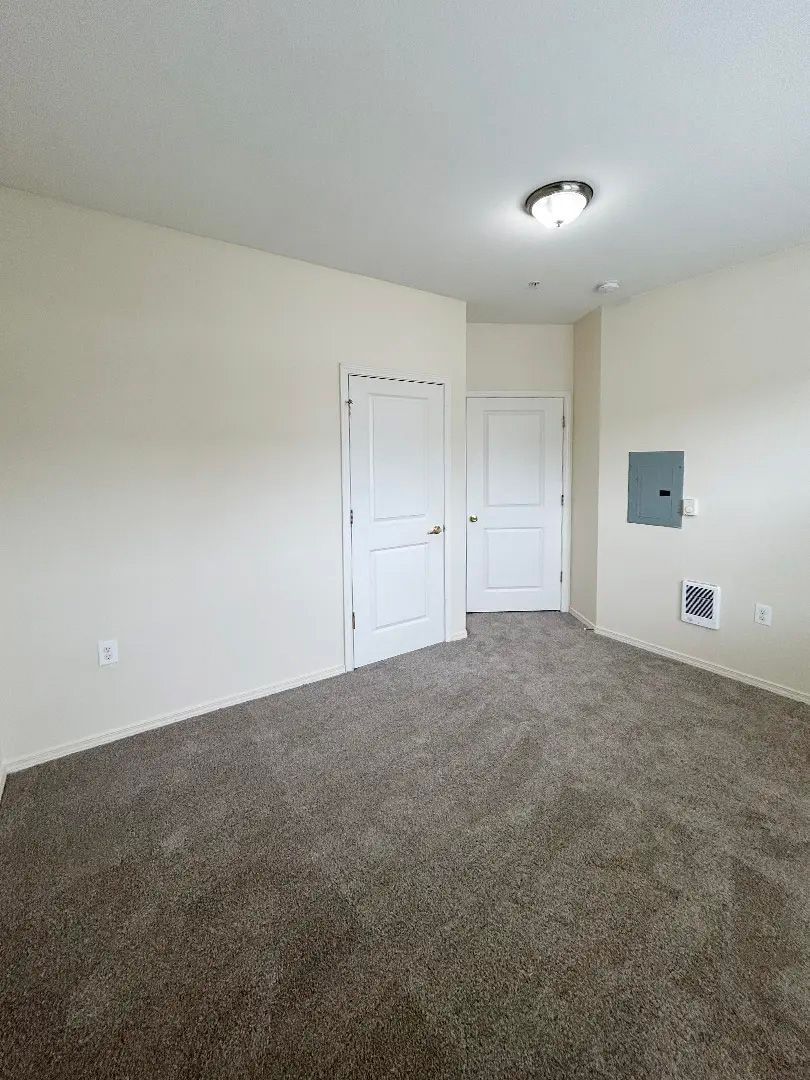 Empty beige bedroom with carpet, white doors, ceiling light, and electrical panels on the wall