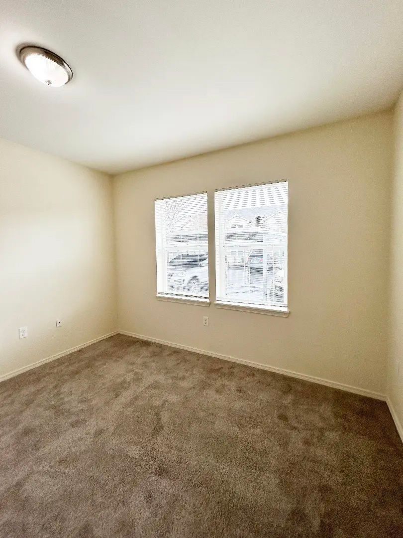 Empty beige bedroom with carpet, white ceiling light, and a window with blinds