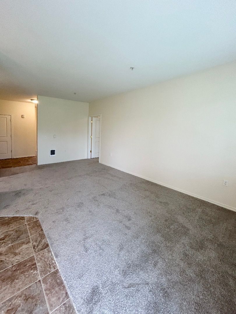 Empty beige living room with gray carpet and tiled entryway.