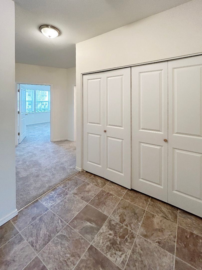 Hallway with tiled floor, white closet doors, and a view into a bright carpeted room