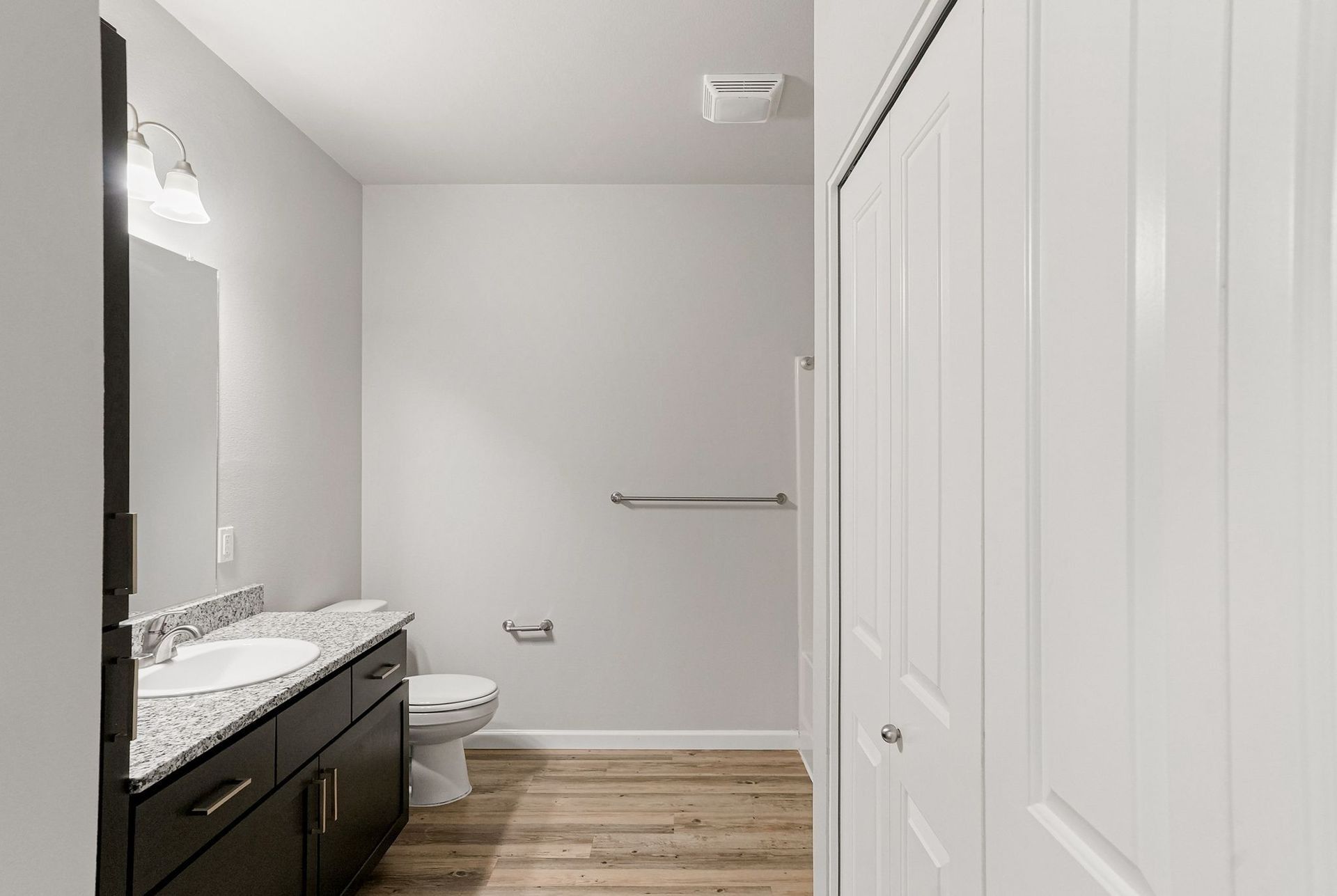 Modern bathroom with white walls, dark vanity, toilet, towel bar, and closet doors on wood-look flooring