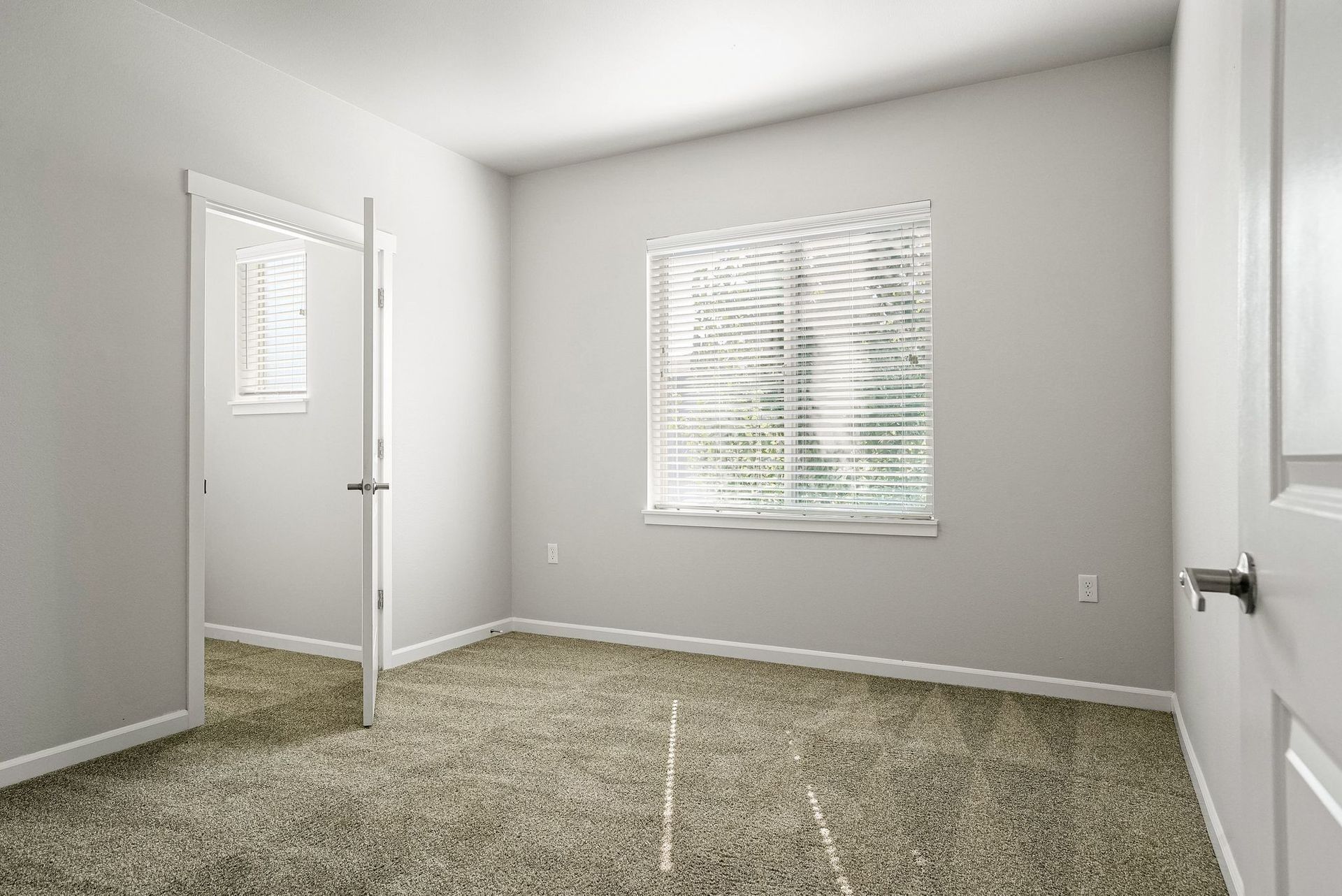 Empty beige bedroom with carpet, window blinds, mirrored closet doors, and open door