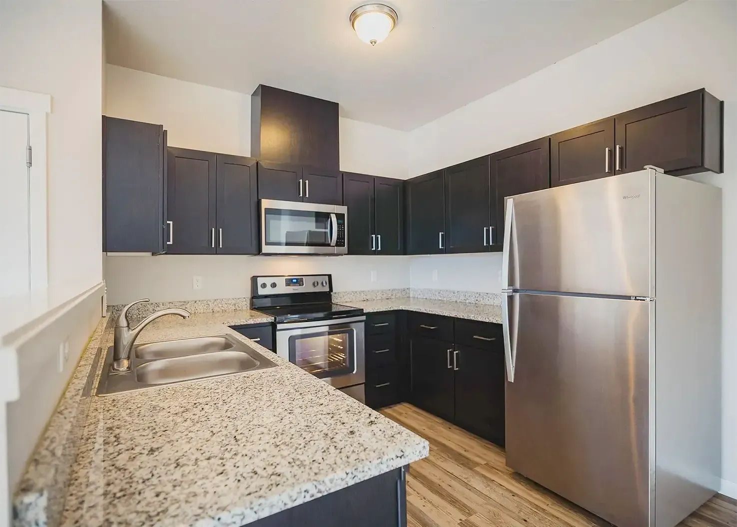 Small modern kitchen with dark cabinets, stainless appliances, white walls, and speckled countertops