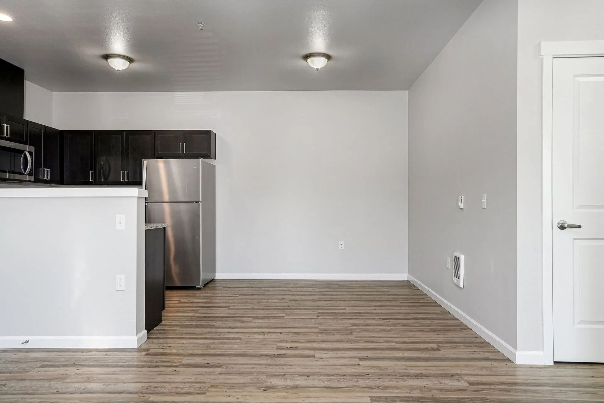Empty modern apartment kitchen with stainless steel fridge, dark cabinets, and light wood floors
