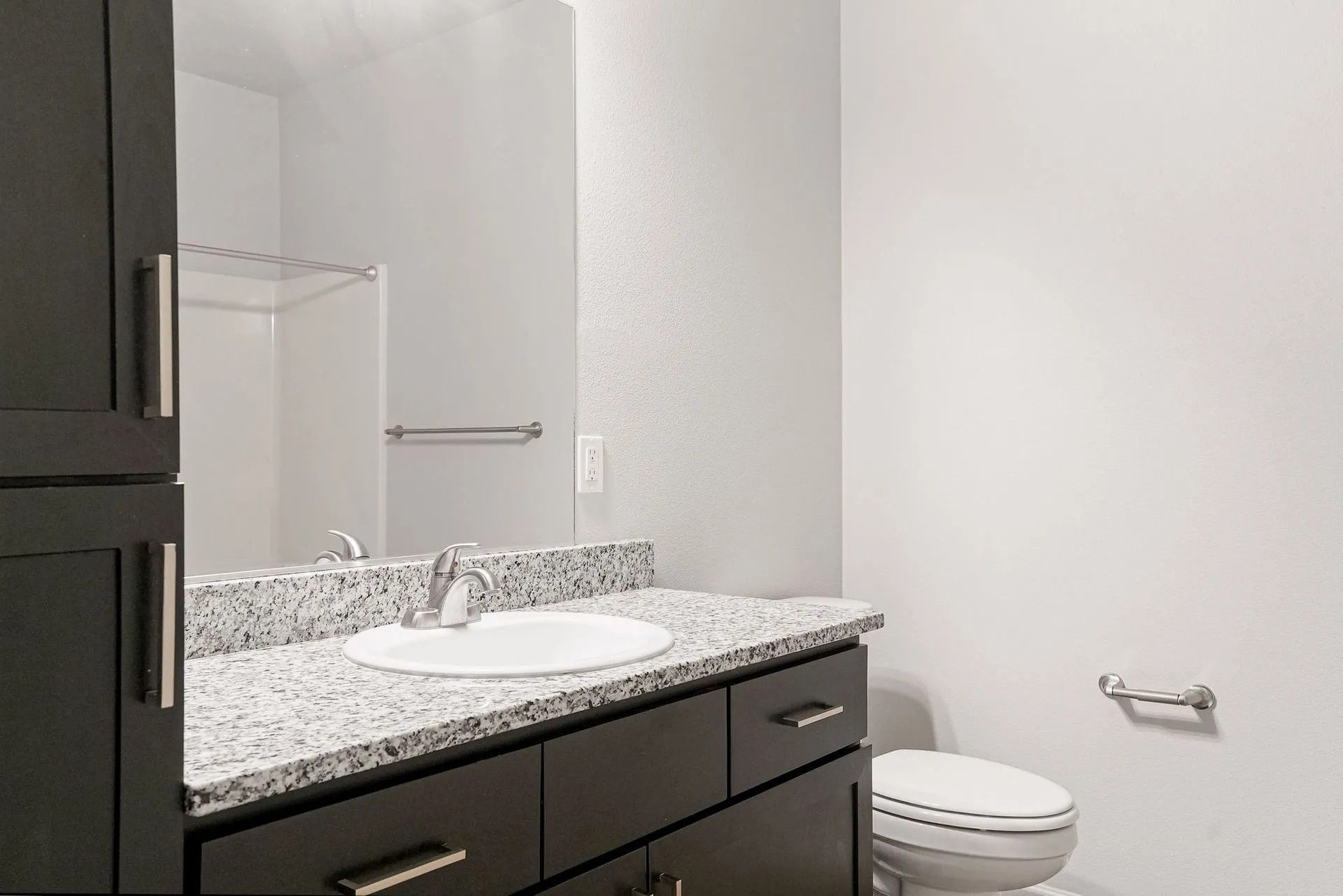 Bathroom vanity with granite countertop, sink, mirror, and toilet against white walls.