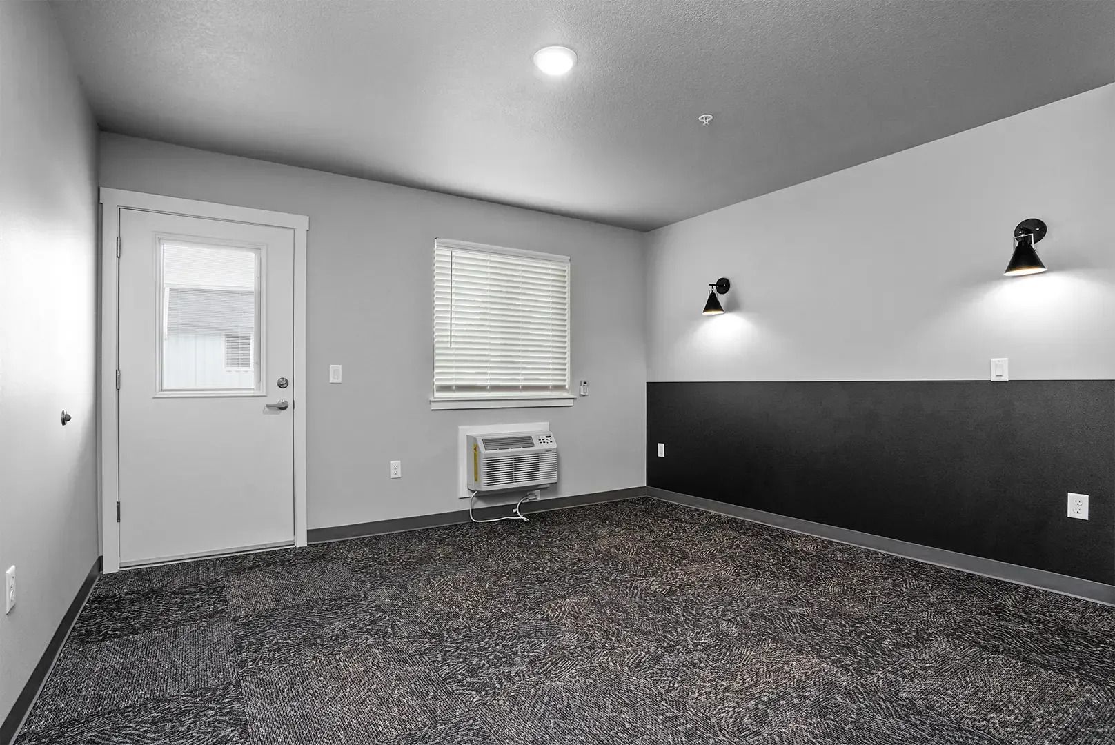 Empty gray carpeted room with a white door, small window, and wall-mounted lights on the black accent wall
