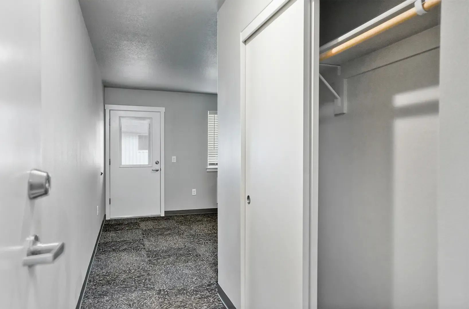 Empty gray hallway with open closet and carpeted floor leading to a door at the end