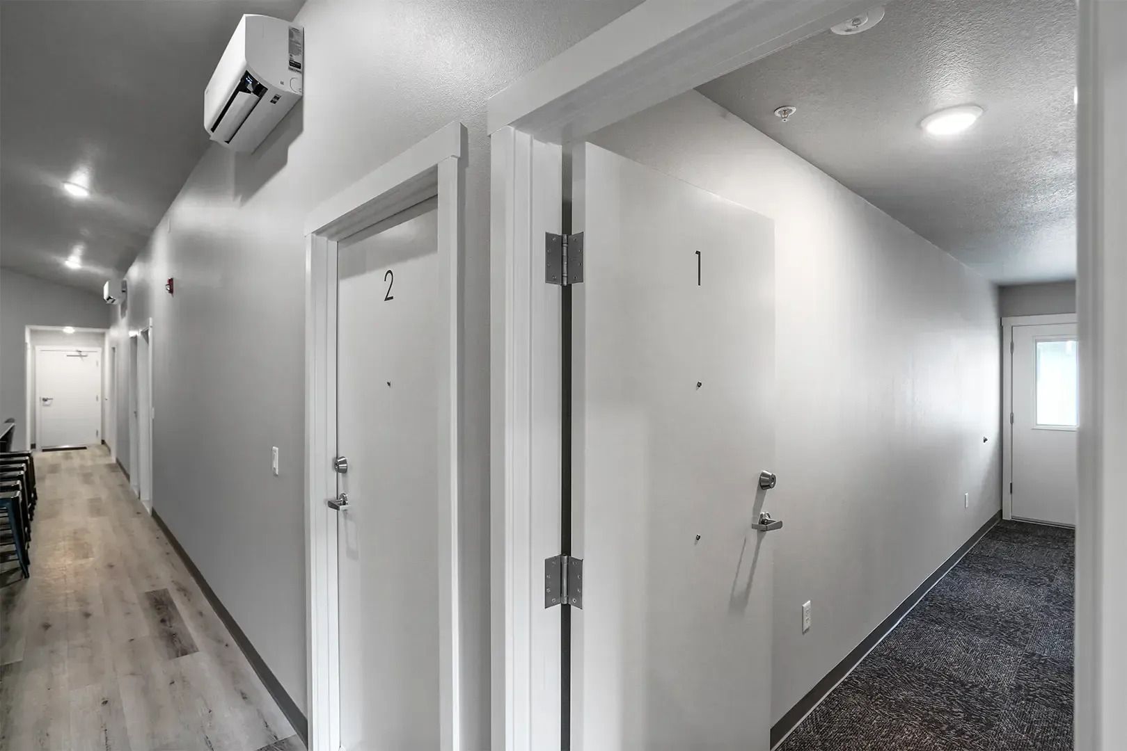 Bright apartment hallway with white doors, wood flooring, and a carpeted corridor turning right.