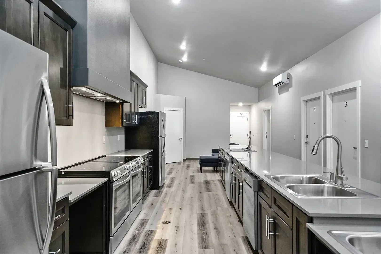 Modern galley kitchen with stainless steel appliances, dark cabinets, white walls, and a long island with sink.