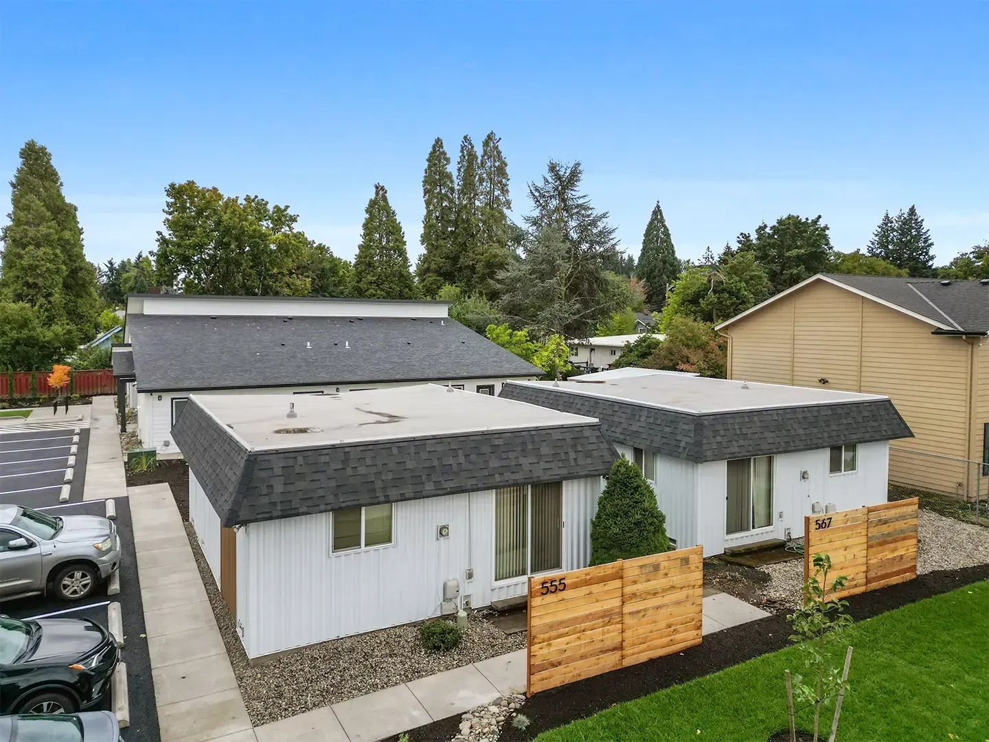 Modern white single-story house with gray roofs, fenced front yard, and trees in the background