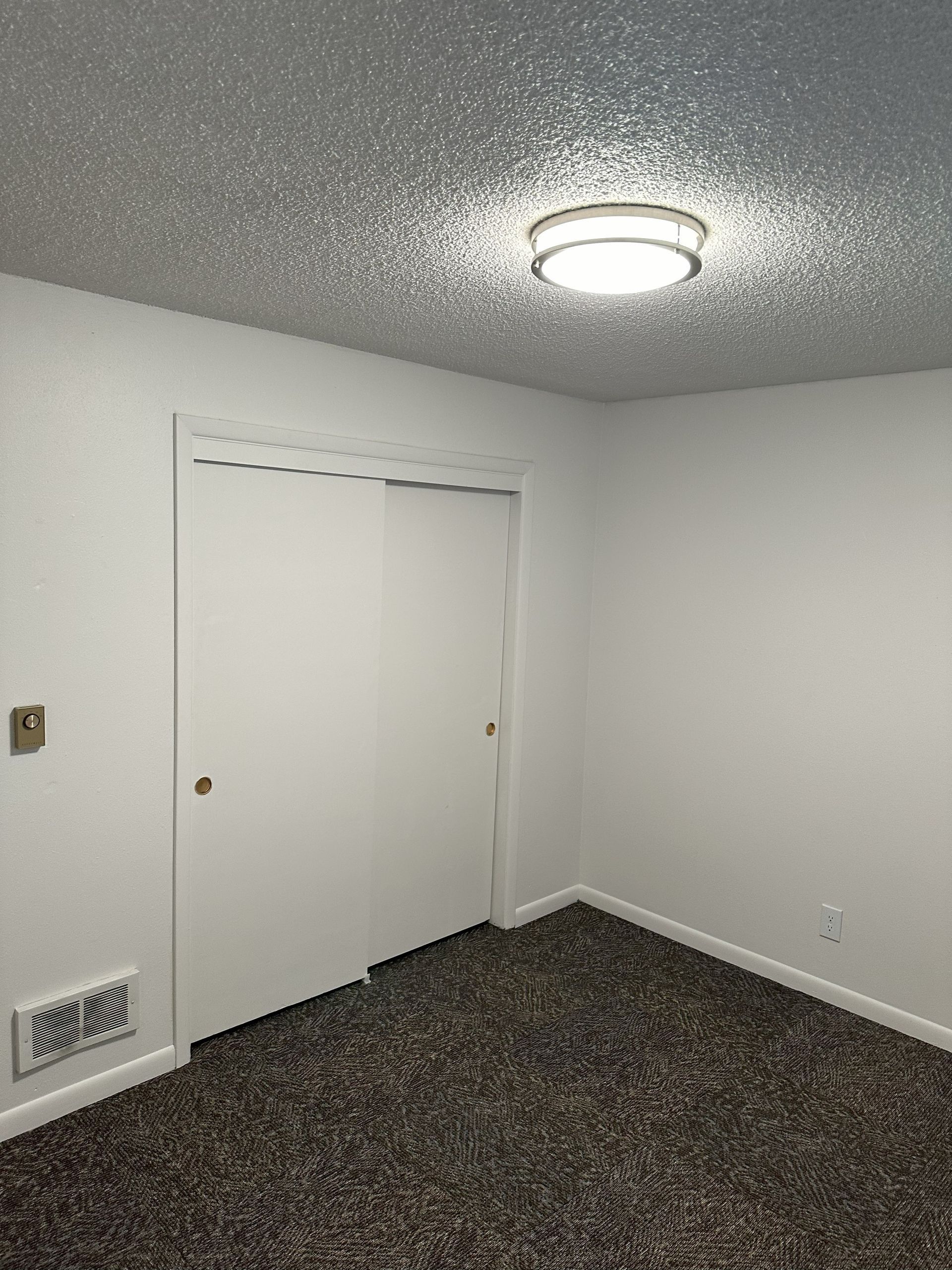 Empty white room with closed closet doors, textured ceiling light, and dark carpet.