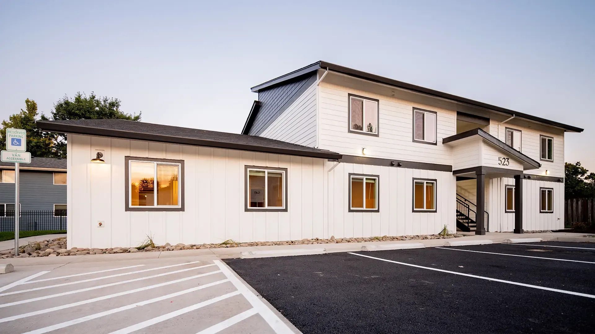 Two-story white house with lit windows and a front parking area at dusk