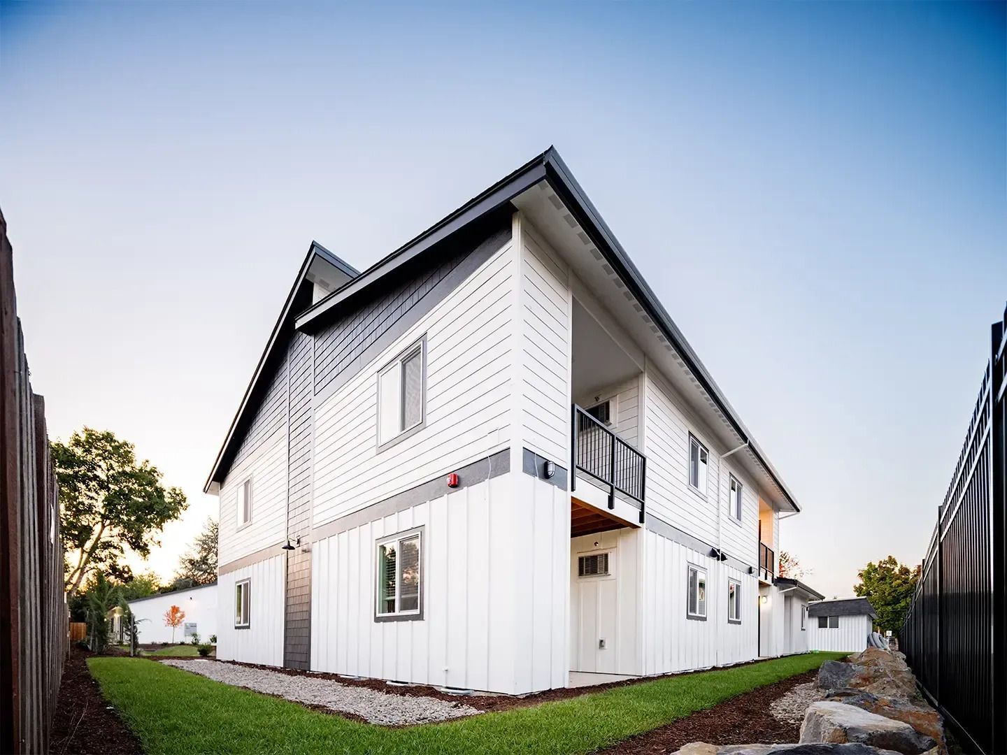 White two-story house with black roof and balcony, viewed from a low angle beside a fenced yard.