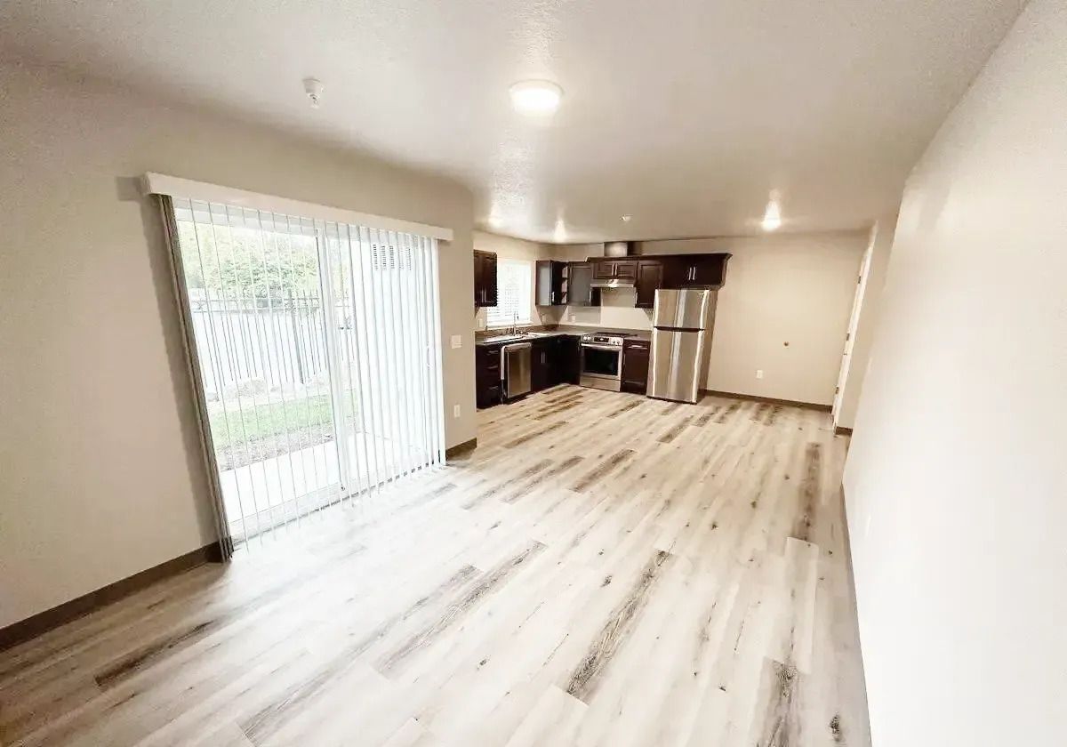 Empty modern living room with light wood floors, sliding glass door, and open kitchen area
