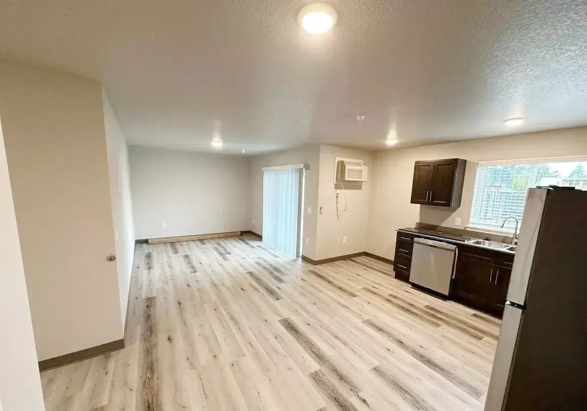 Empty open-plan kitchen and living room with light wood floors, white walls, dark cabinets, and sliding glass doors.