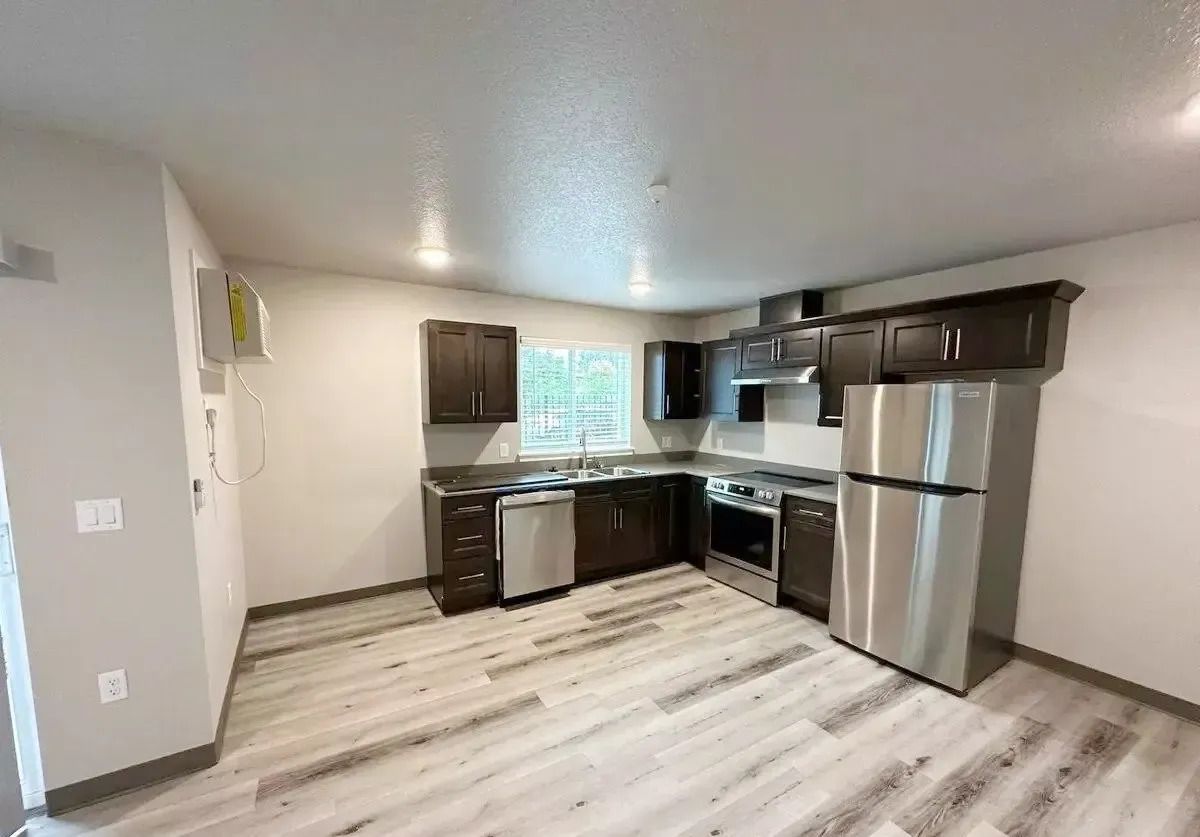 Empty kitchen with dark cabinets, stainless steel appliances, and light wood flooring.
