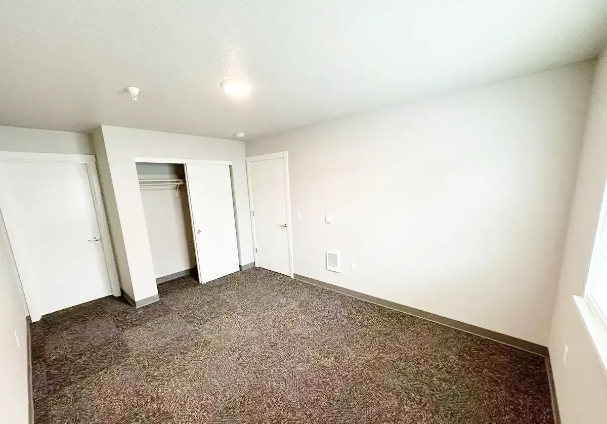 Empty carpeted bedroom with white walls, closet doors, and a window on the right.