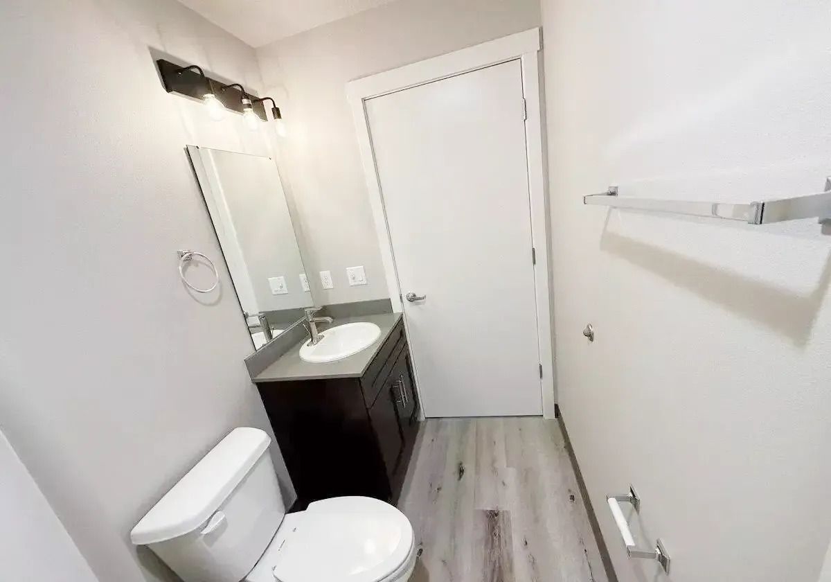 Small bathroom with toilet, vanity sink, mirror, towel bar, and closed door in a narrow white room.