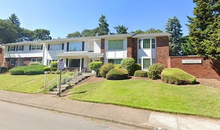 Suburban brick apartment building with green lawn, steps, and parked car on a sunny day