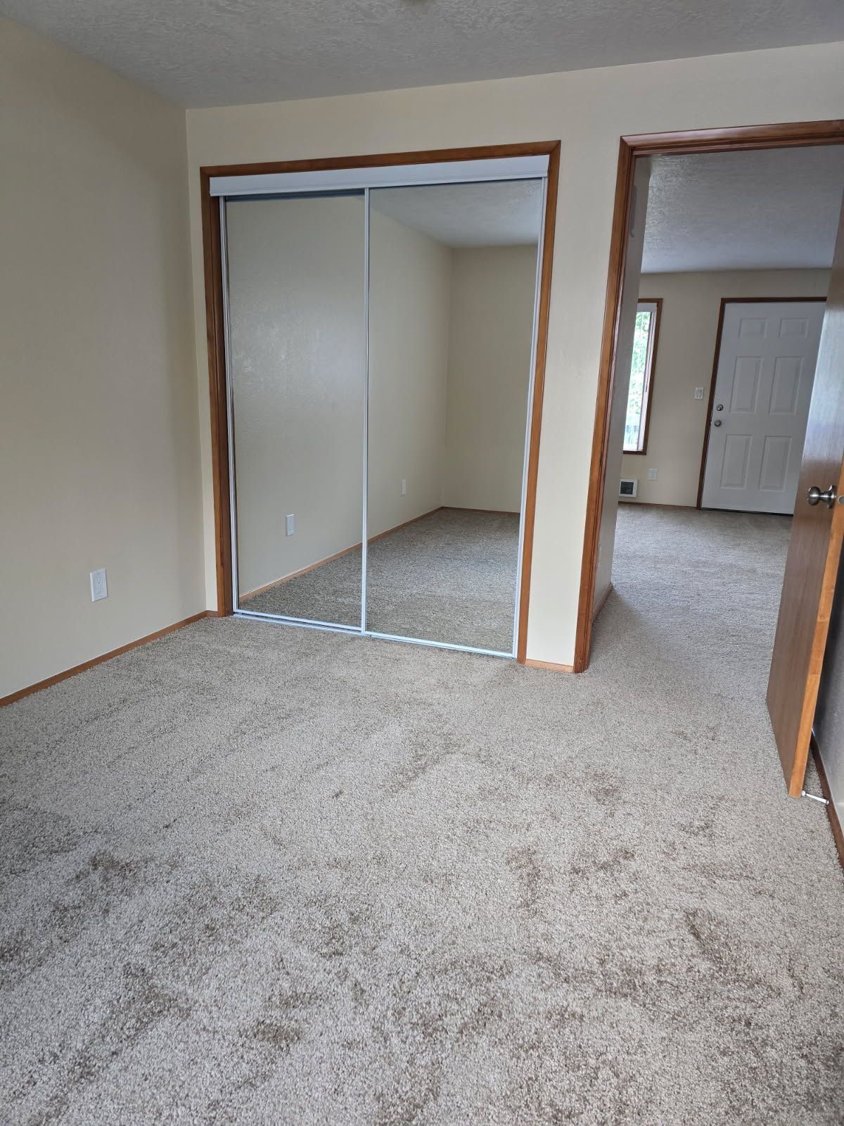 Empty beige bedroom with carpet, mirrored closet doors, and a doorway to another room