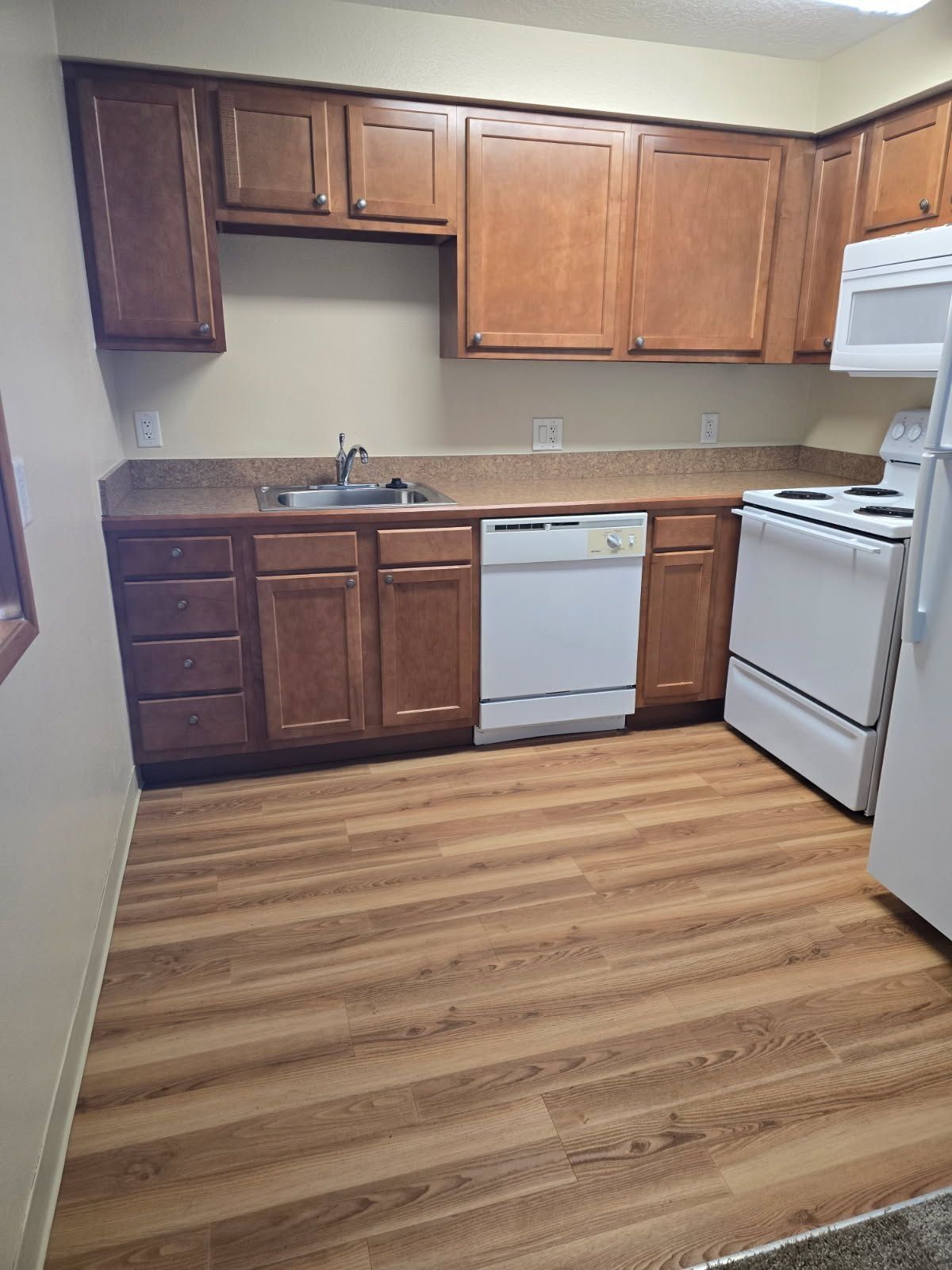 Empty kitchen with brown cabinets, white appliances, and wood-look flooring.