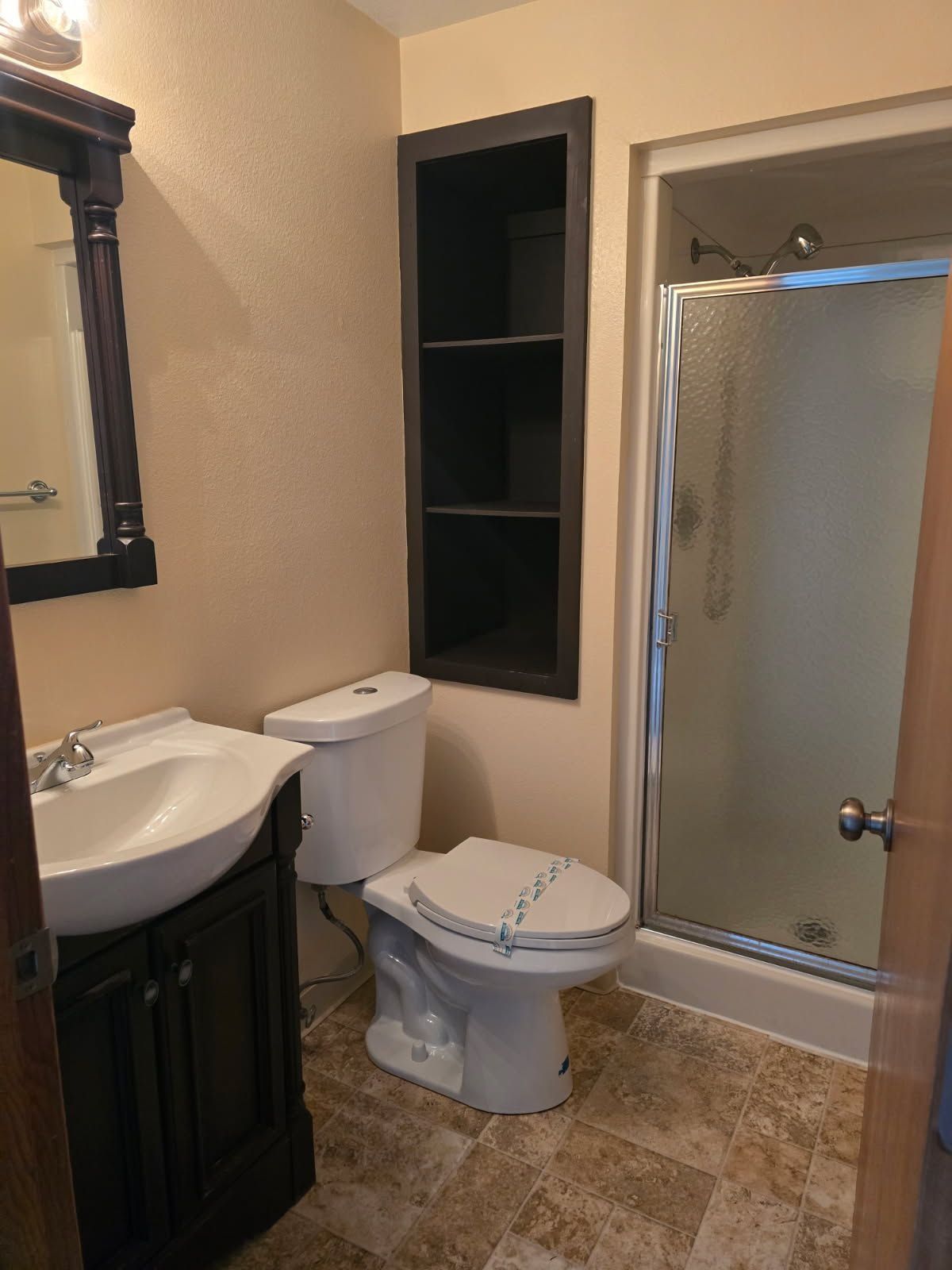 Small beige bathroom with toilet, sink vanity, shower, and wall mirror.