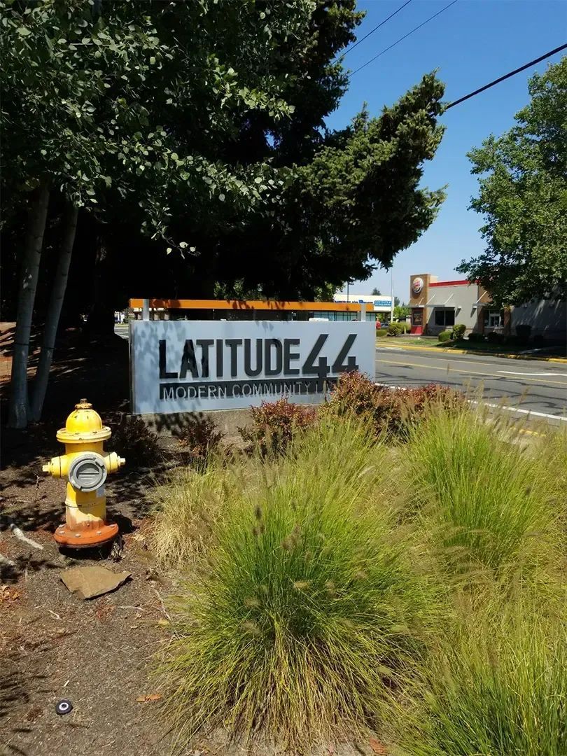 Latitude 44 roadside sign beside a yellow fire hydrant, with tall grass and trees on a sunny street