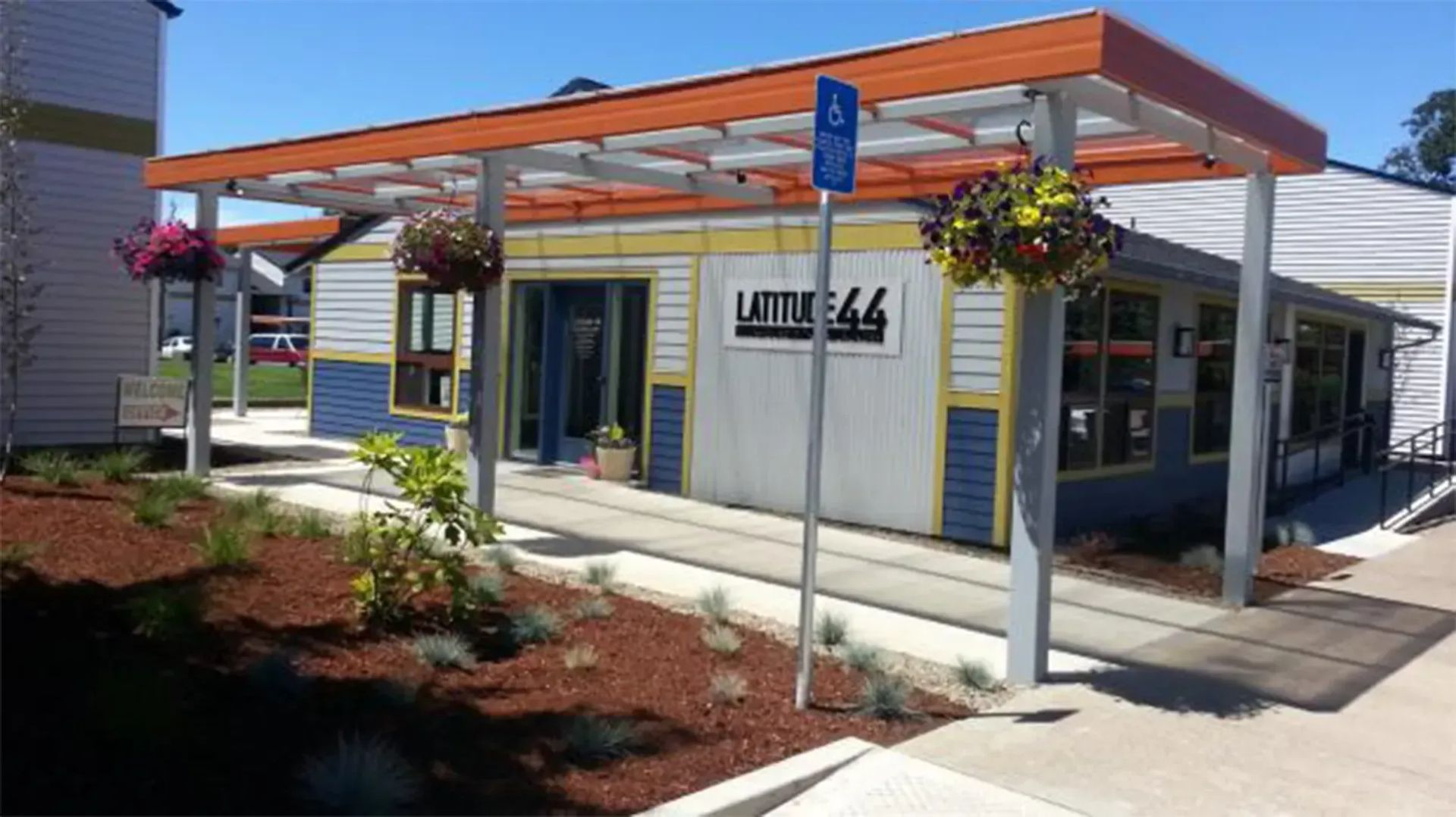 Modern storefront with orange awning, hanging flowers, and blue-trimmed entrance in a shopping plaza