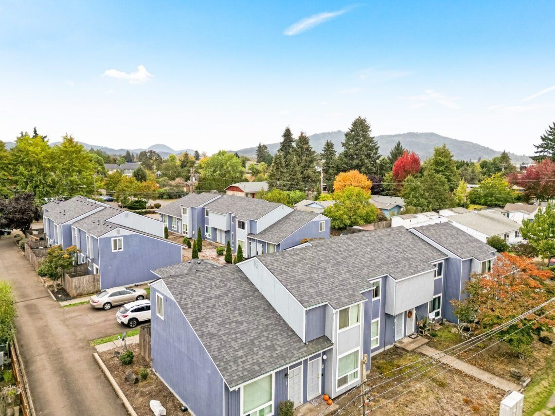 Aerial view of suburban townhouses with gray roofs, tree-lined streets, and distant hills under a clear sky