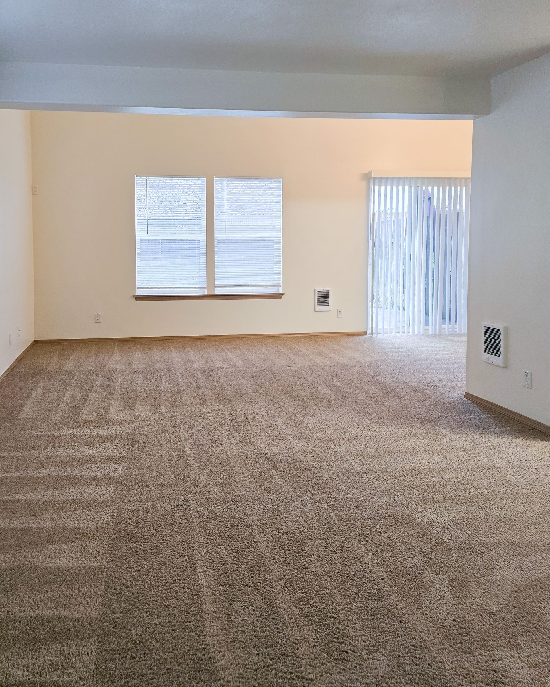 Empty carpeted living room with beige walls, two windows, and a sliding glass door with vertical blinds